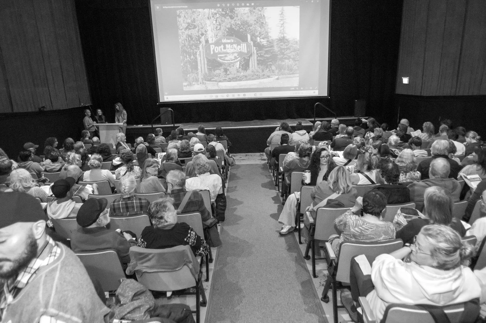 A large audience seated in a theater or conference room, watching a presentation on a large screen with an image of a sign that reads 'Welcome to Port McNeill'.