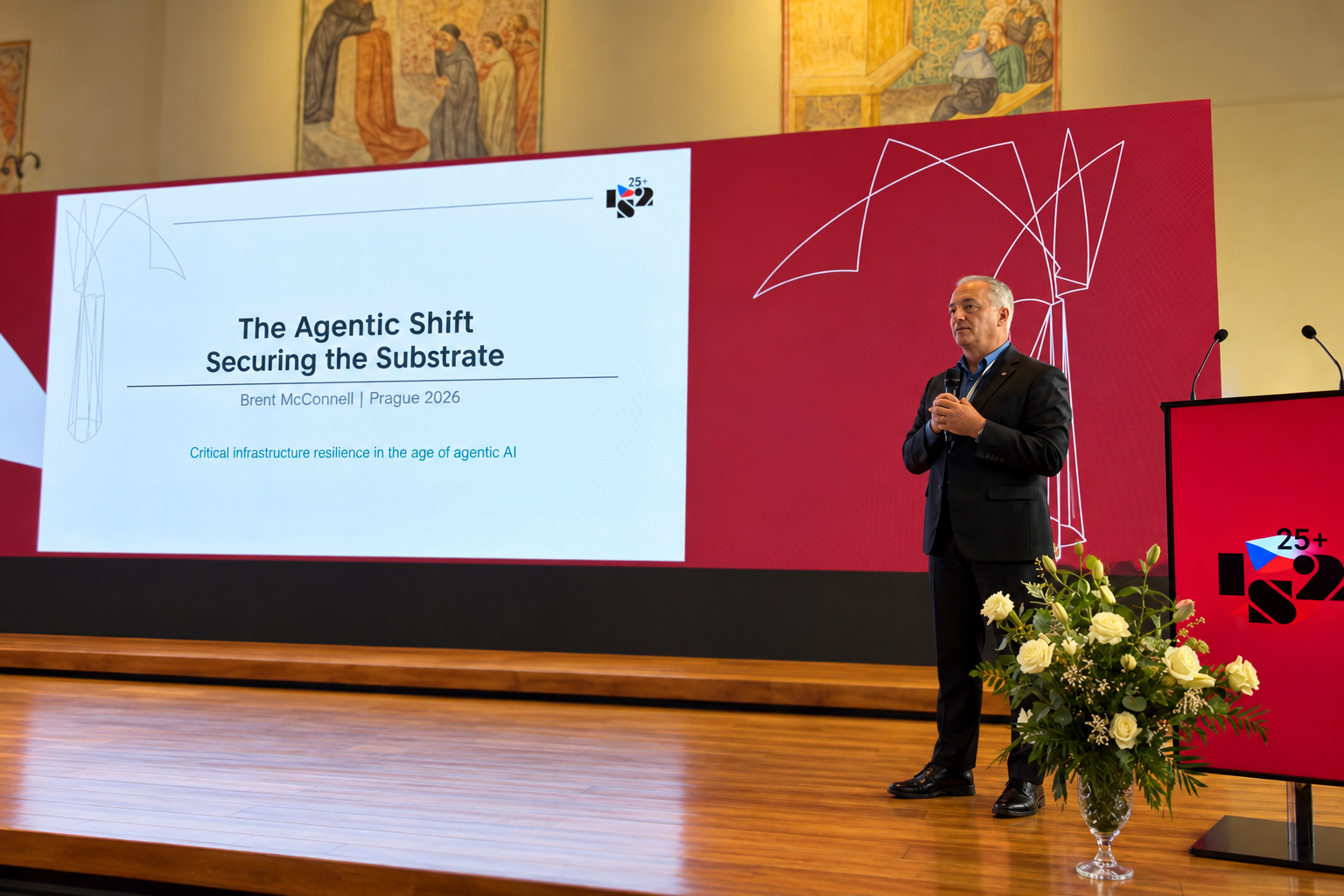 A man in a dark suit holding a microphone stands on a wooden stage next to a large presentation screen and a red podium with a stylized logo. The presentation slide reads: 'The Agentic Shift Securing the Substrate, Brent McConnell, Prague 2026, Critical infrastructure resilience in the age of agentic AI.' There is a bouquet of white roses and greenery on the stage, and the background features historic religious or medieval artwork.
