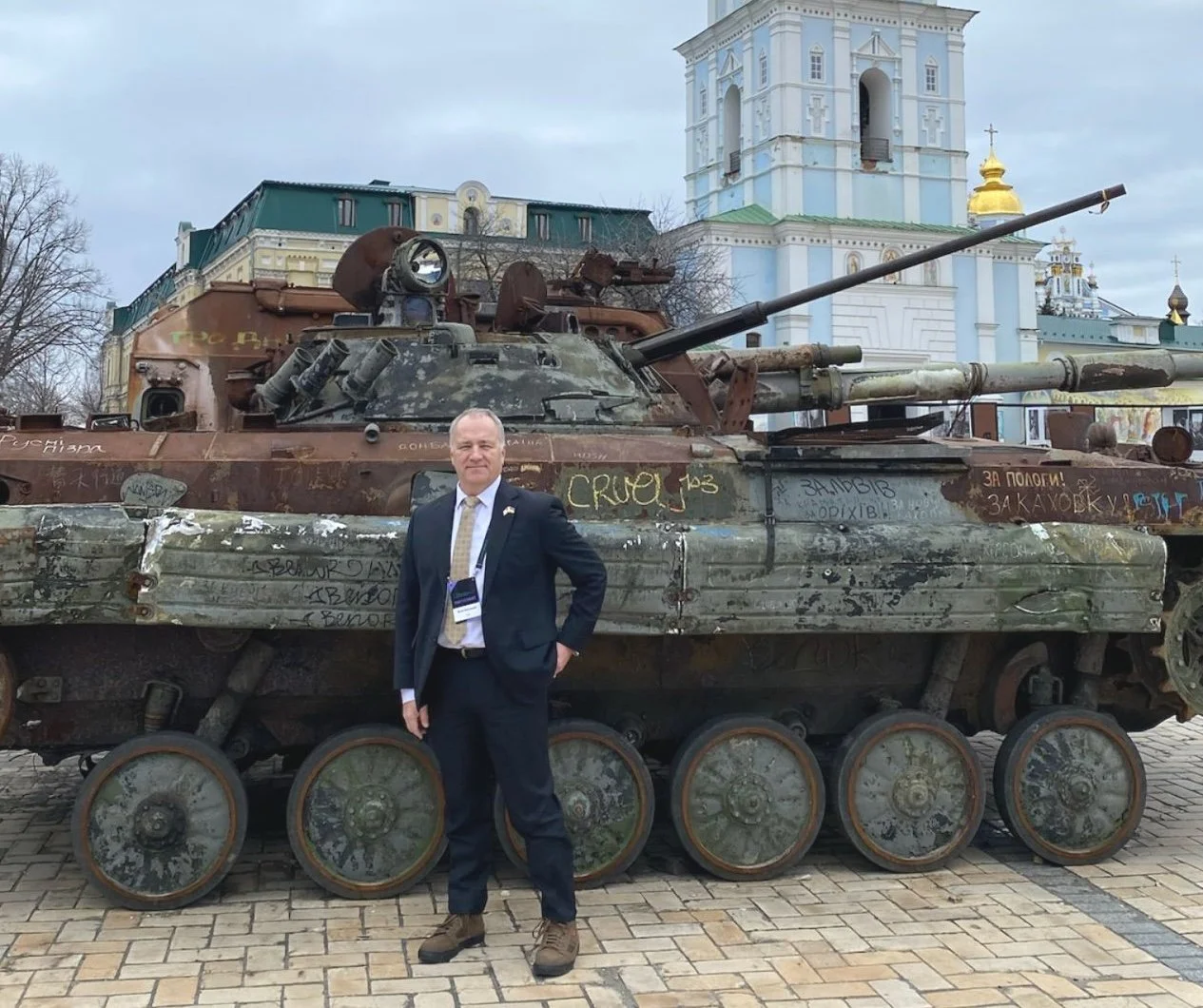 A man in a suit standing in front of a rusty, graffitied tank in Ukraine on a city street, with historic buildings and church towers in the background.
