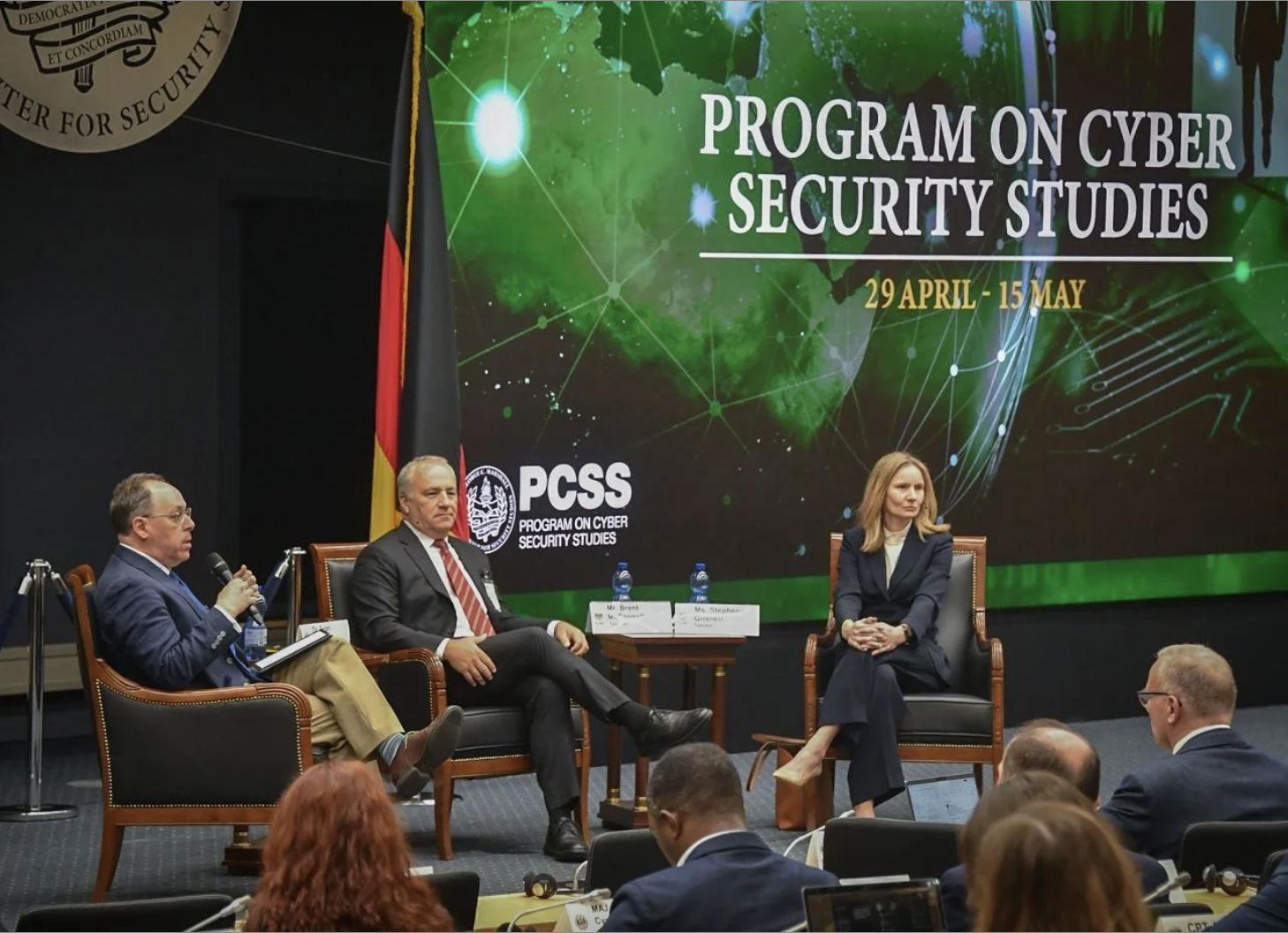 Panel discussion at a conference on cyber security studies with three speakers seated on stage, a large screen behind showing the event title and dates, an emblem, and a German flag.