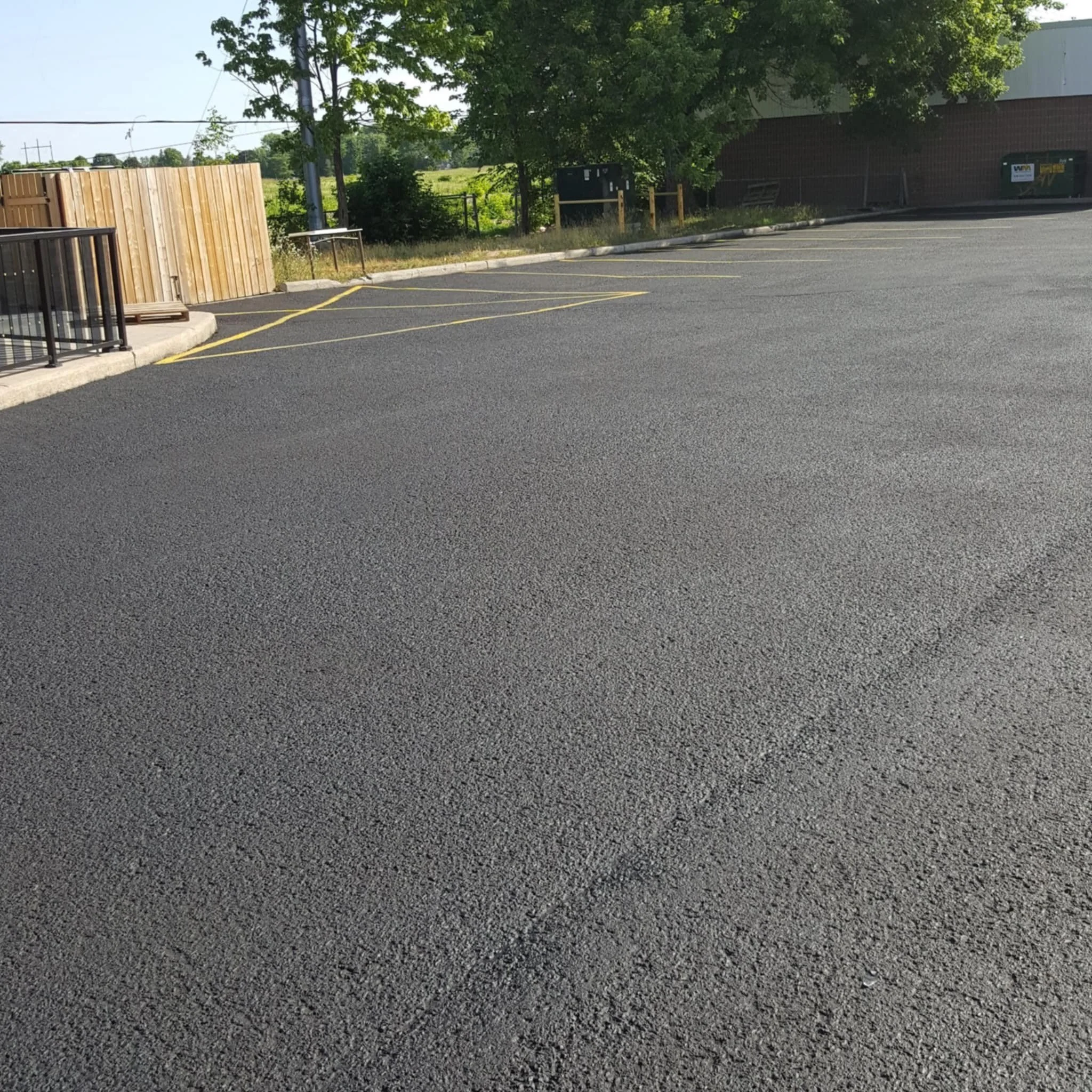 Empty parking lot with freshly paved black asphalt, yellow parking lines, a tree providing shade, and a building with a brick exterior in the background.