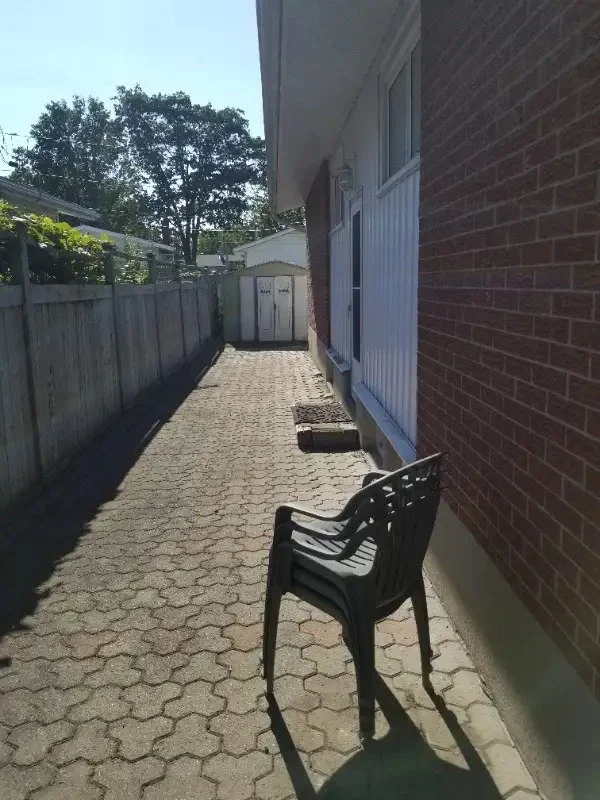 A narrow paved outdoor walkway beside a brick house with a white window and a black plastic chair, leading to a small shed at the end.