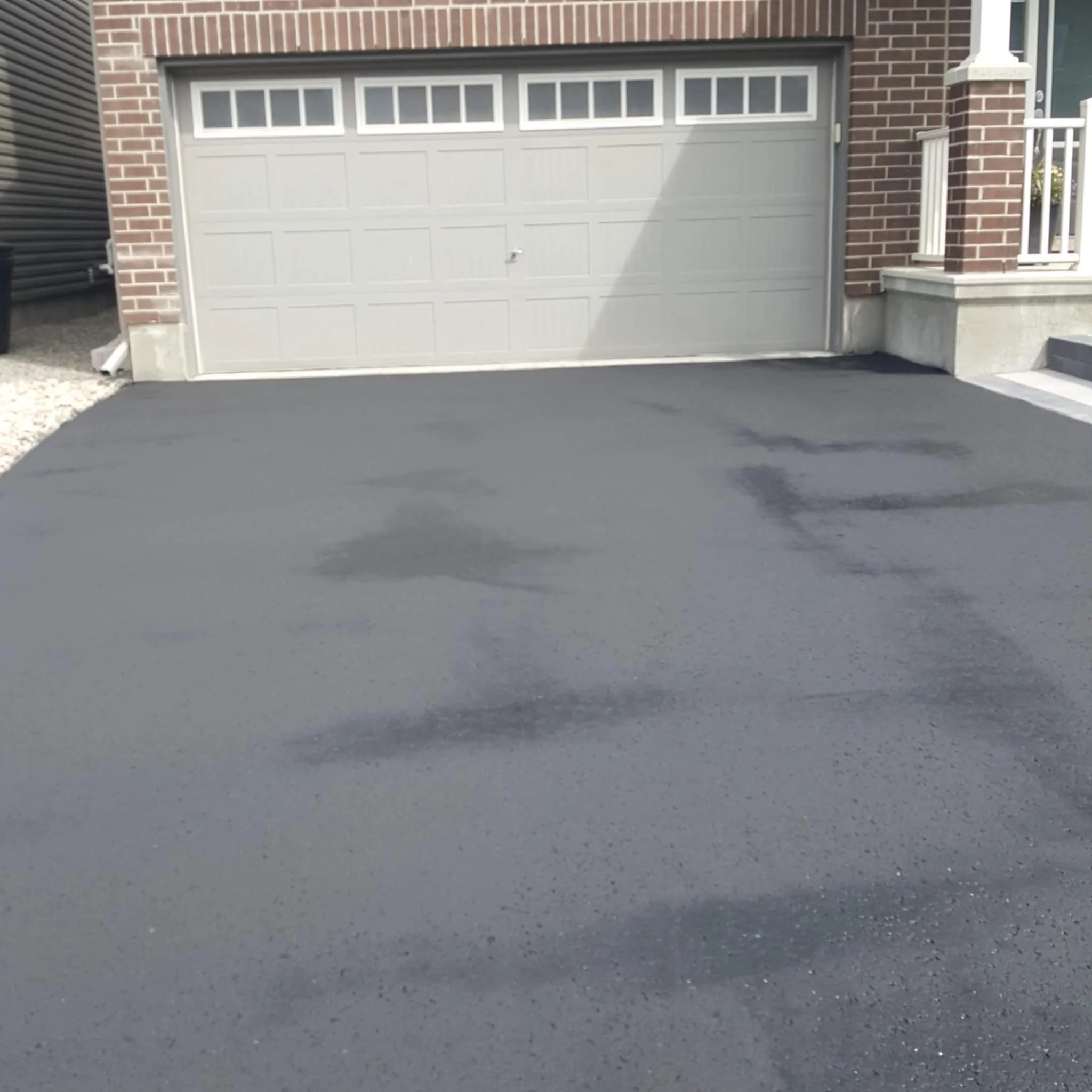 Freshly paved black asphalt driveway in front of a white garage door attached to a brick house.