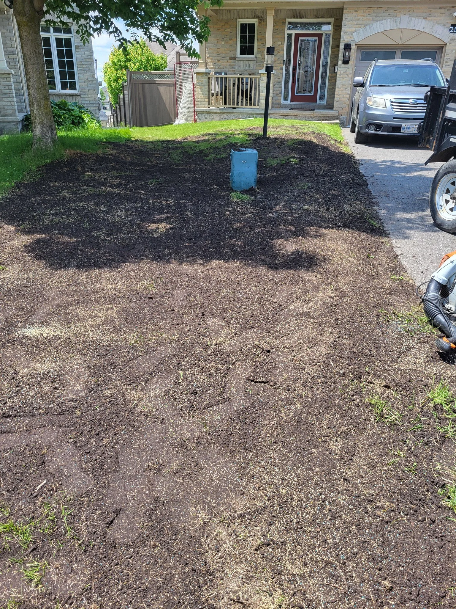 A yard with freshly turned and disturbed soil, some green grass patches, and a new mulch layer, with a house, driveway, and parked vehicles in the background.