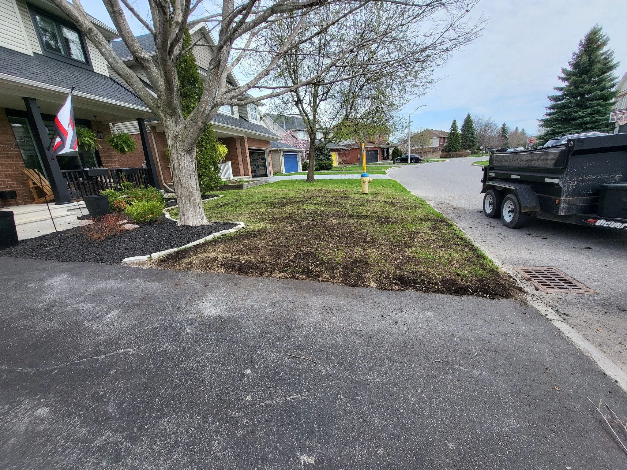 A residential street with a front yard being landscaped, showing a tree, patches of dirt, a barbecue trailer, and a house with a porch and a flag.