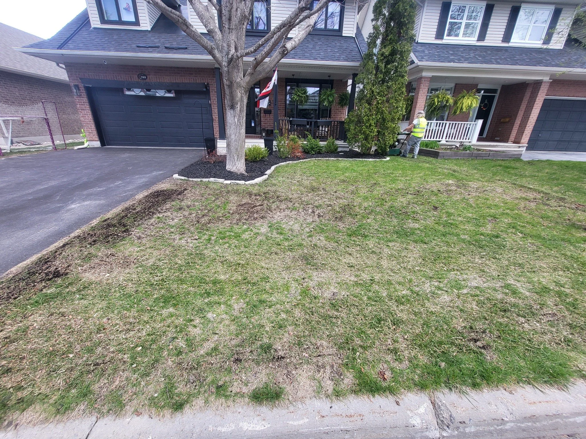 Front yard of a house with a tree, some shrubs, and a worker trimming a bush. The house has a black garage door with two small windows, brick exterior, and a porch with hanging plants.