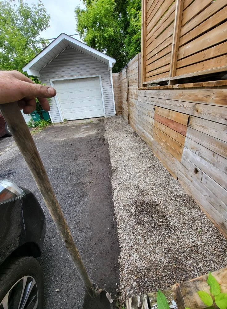 Person using a shovel to spread gravel along the edge of a driveway beside a wooden fence, with a garage in the background.