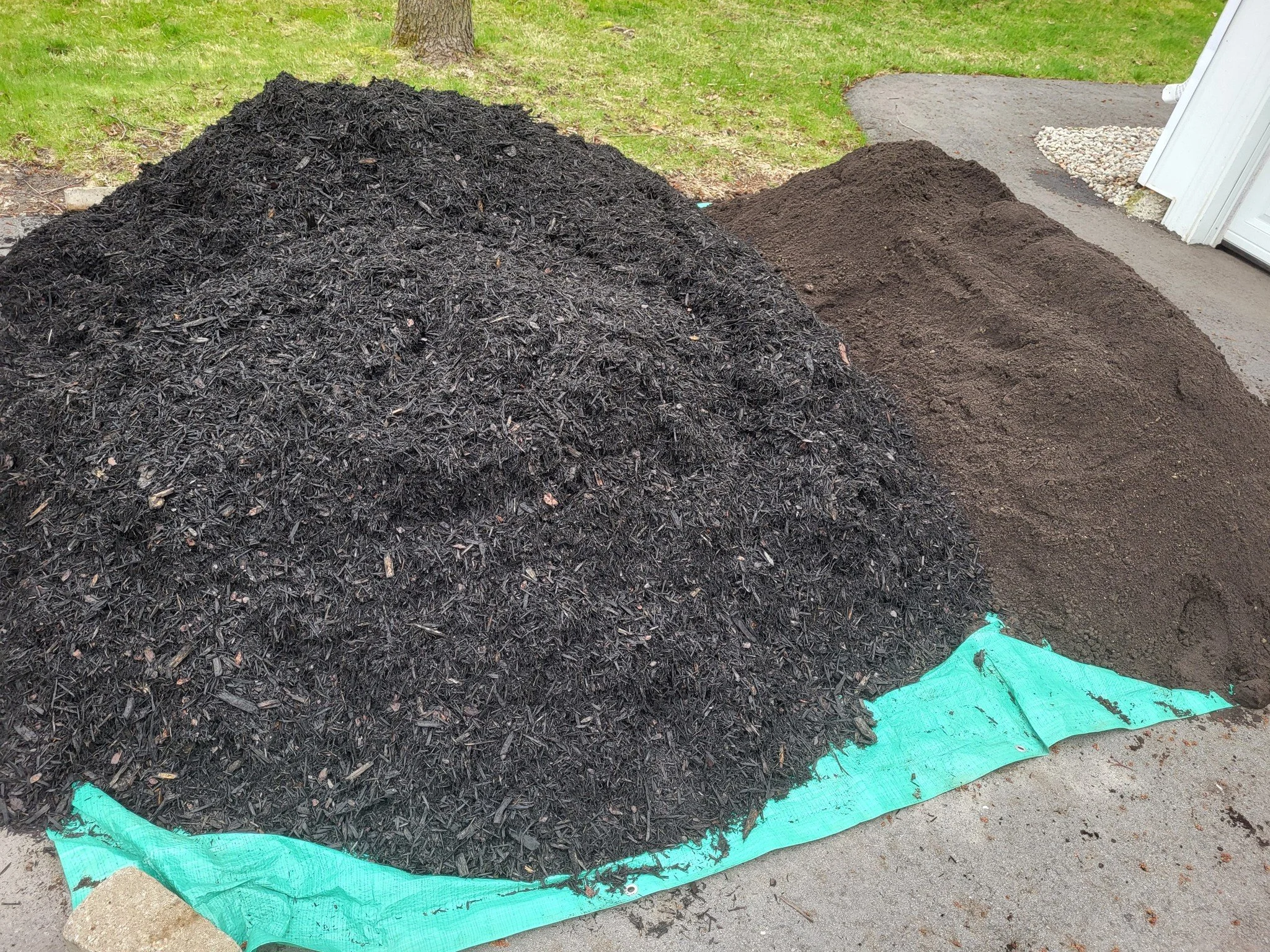 Two piles of gardening materials on a concrete driveway, one of composted mulch and the other of dark soil, with a green tarp partially visible beneath them.