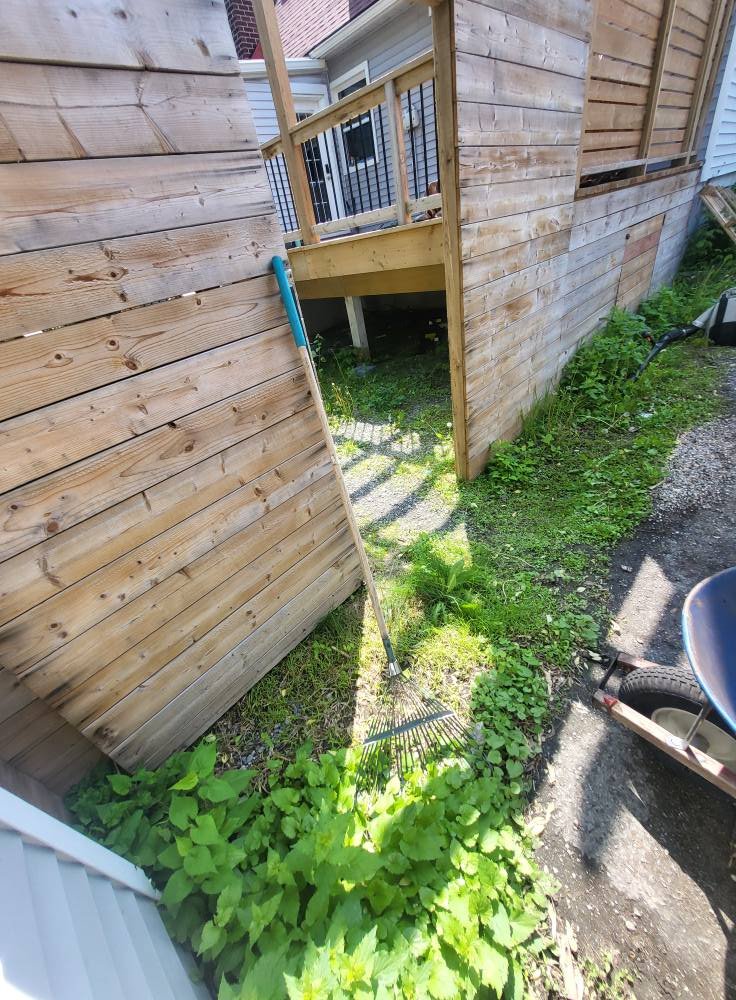 A small construction project shows a partially built wooden deck with a black metal railing attached to a house. There's a green garden rake with a blue handle leaning against a wooden fence, and some green plants growing near the ground. A wheelbarr