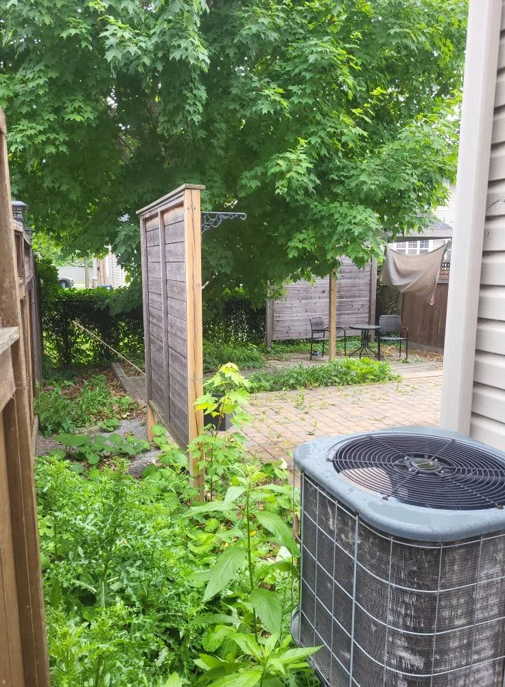 View of a backyard garden with lush green trees, a wooden privacy fence, a small patio area with outdoor furniture, a garden bed with plants, and an air conditioning unit in the foreground.