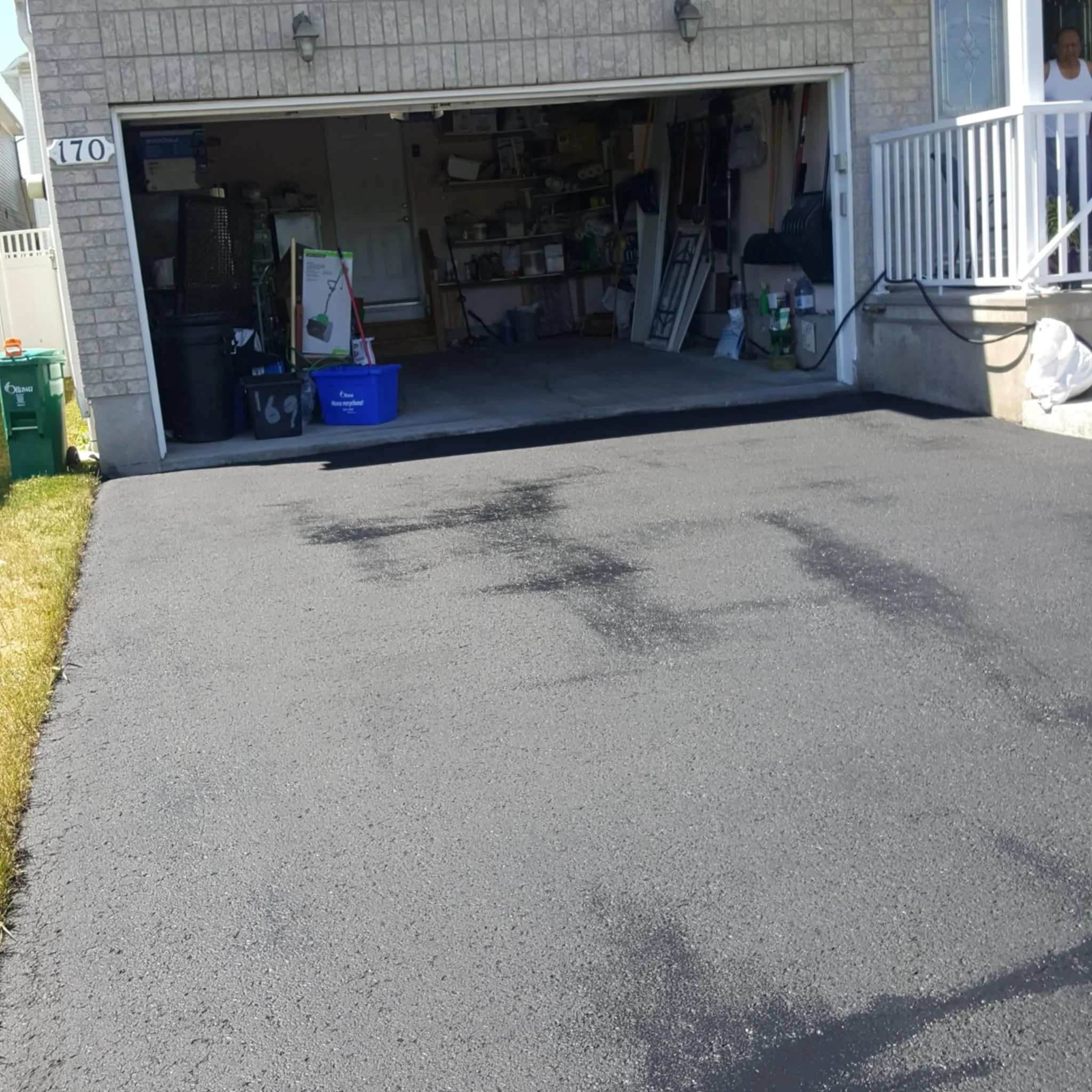 Open garage with tools and storage shelves, driveway with tire tracks, yellow grass on left, house number 170 on brick wall, person partially visible on balcony.