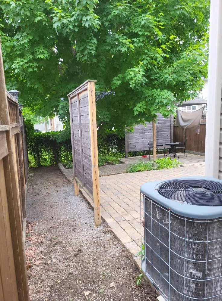 Backyard with a large green tree, a wooden privacy screen, a bench, and an air conditioning unit.