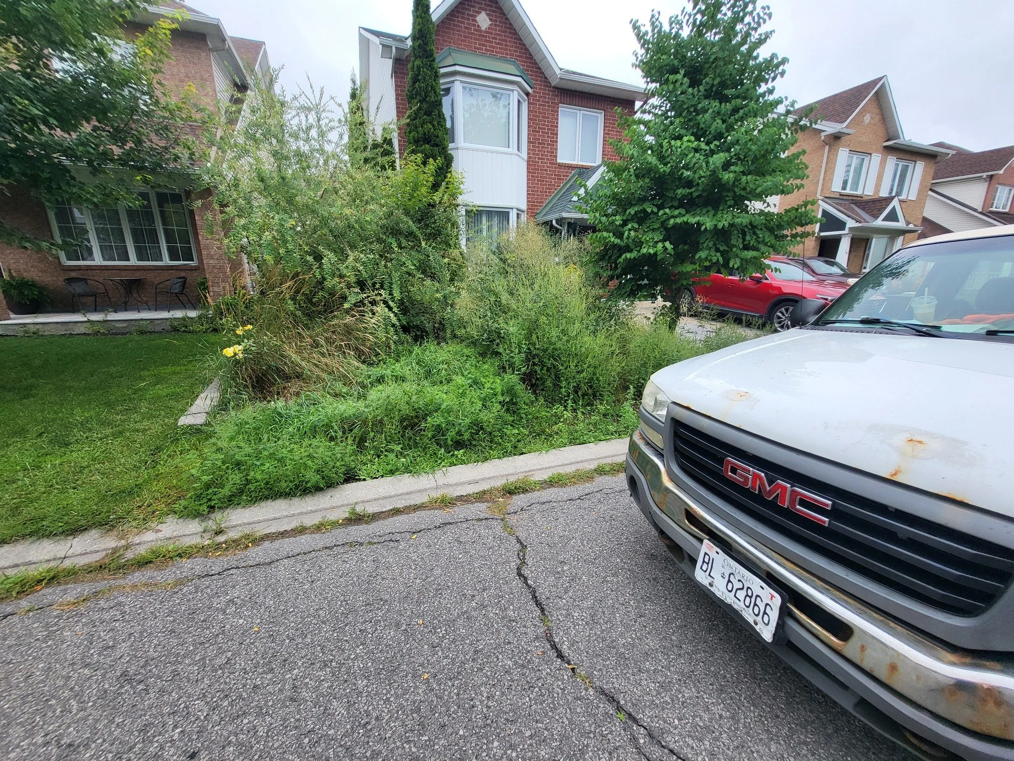 A white GMC vehicle parked on a cracked asphalt street with overgrown grass and bushes alongside a residential sidewalk. In the background, there are two-story brick houses, some with large windows and small porches, on a cloudy day.