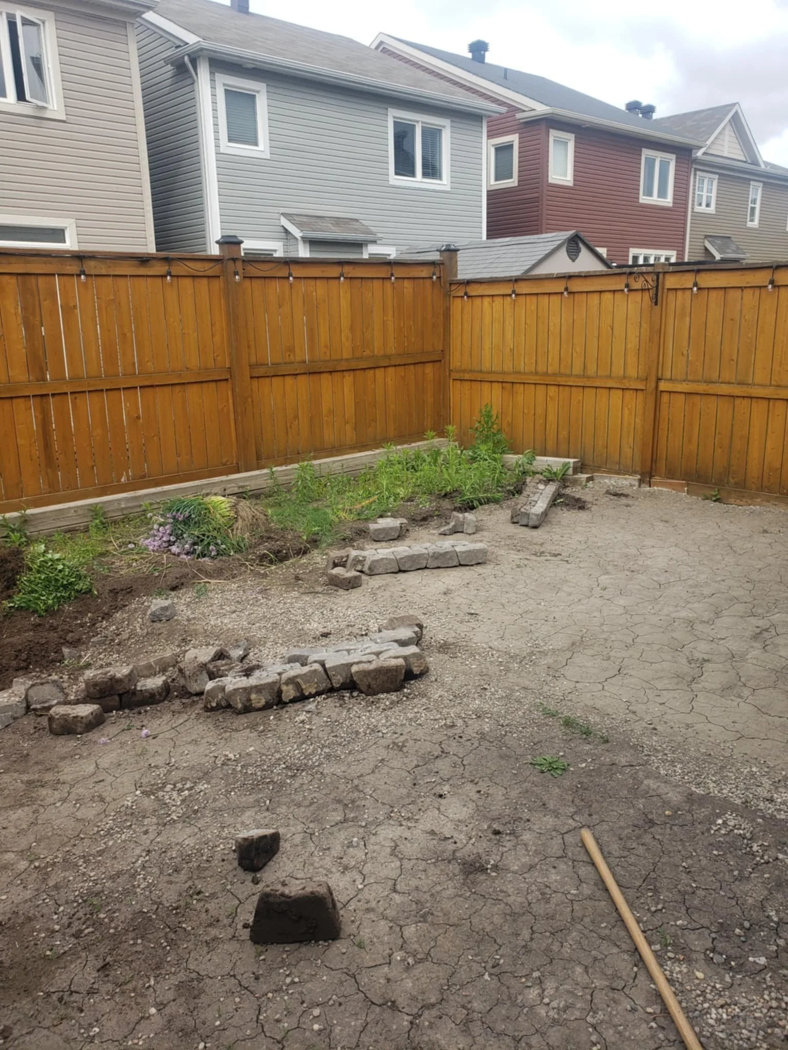Backyard with a wooden fence, plants, and a partially paved area with bricks arranged for a project or garden layout.
