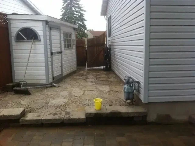 Backyard with uneven stone pathway, small shed on left, house siding on right, yellow bucket in center front, and a closed wooden gate at the back.