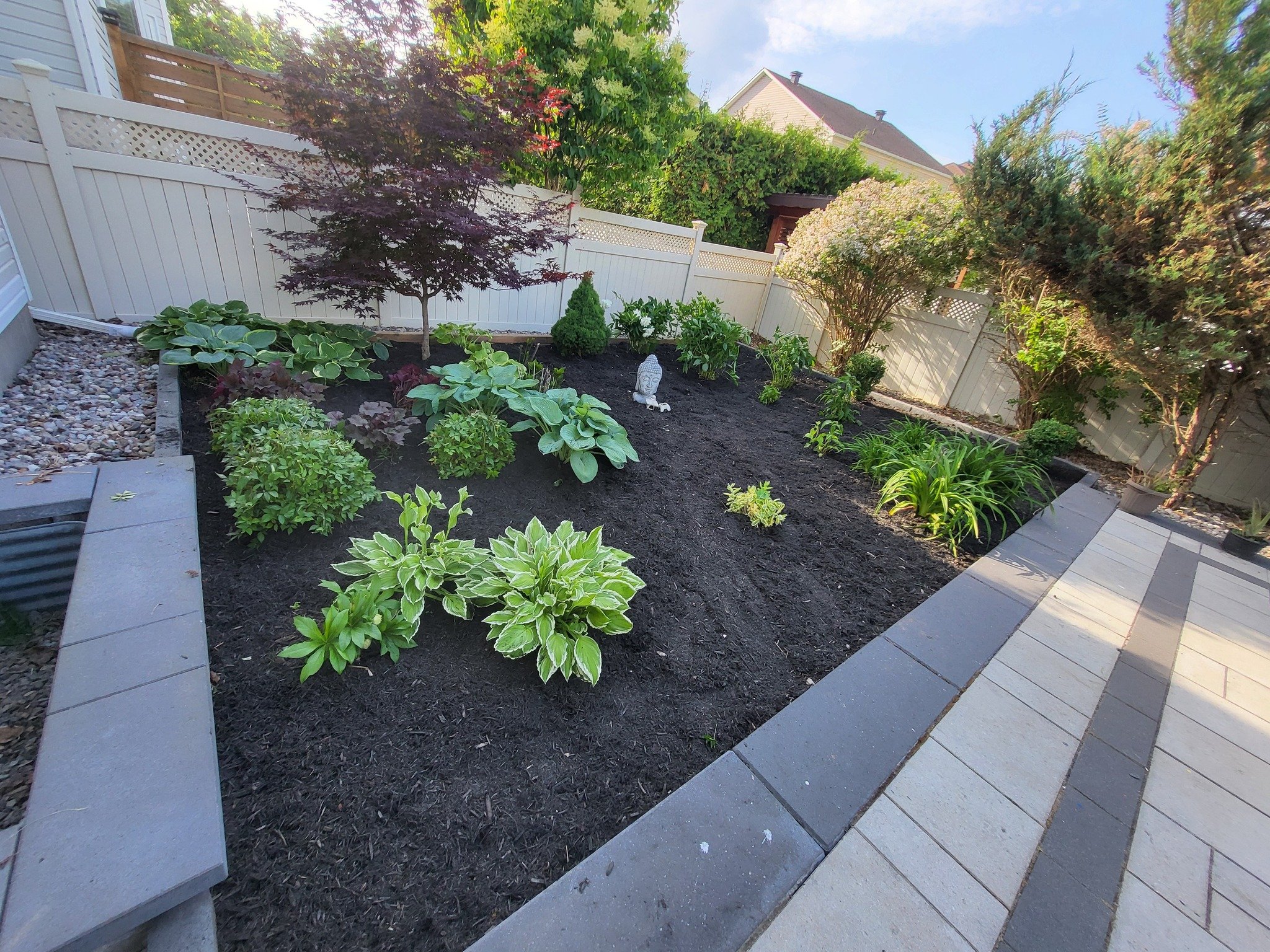 A landscaped backyard garden with various plants, trees, a statue of a Buddha, a white fence, and a paved walkway.