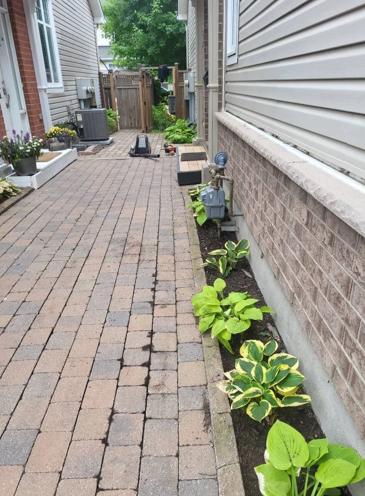 A narrow brick-paved backyard side area with a flower bed along a brick house, a wooden fence at the back, and some gardening tools and plants along the edge.