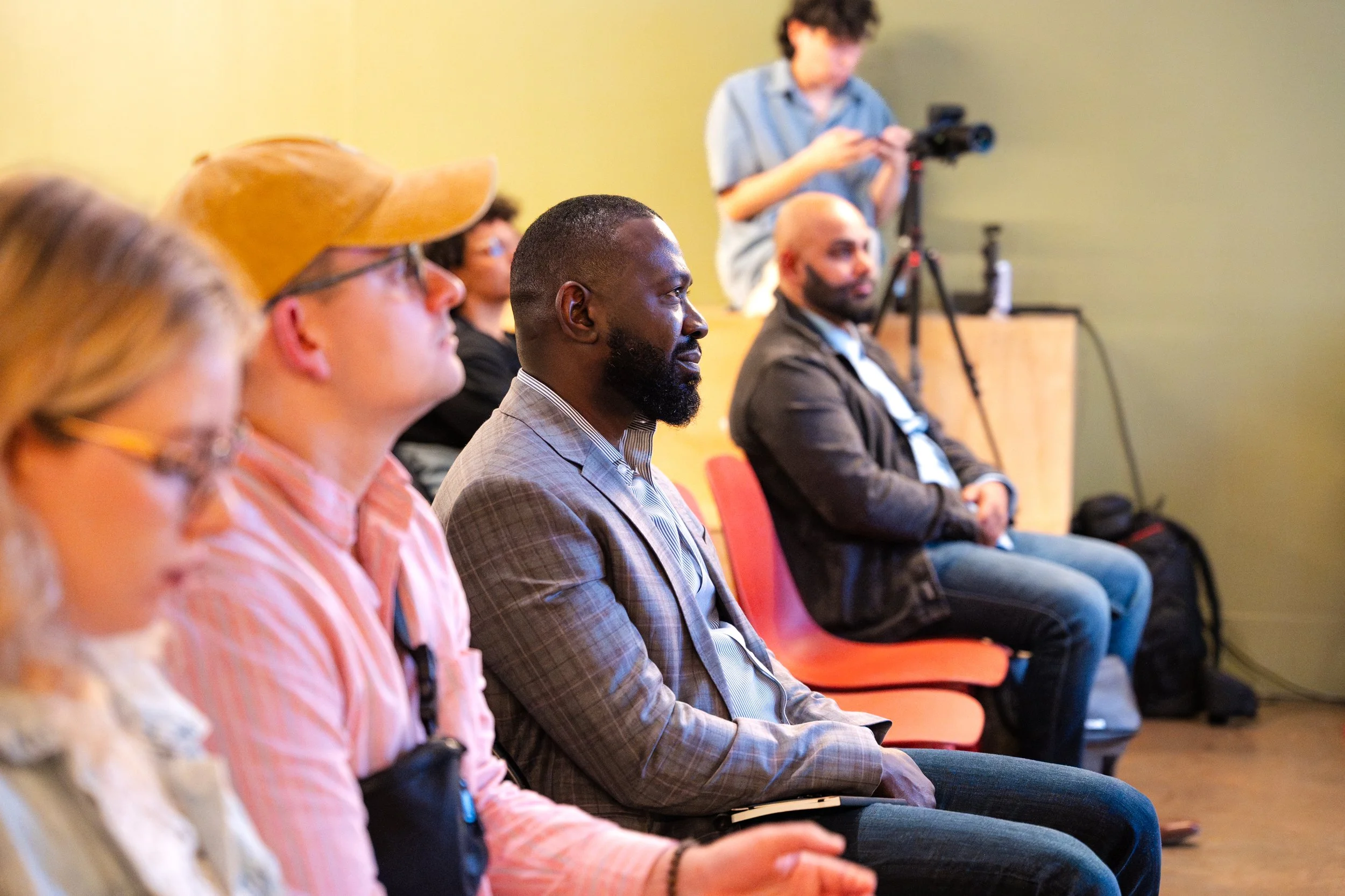 People seated in an audience attending a presentation or conference, with a camera operator filming the event.