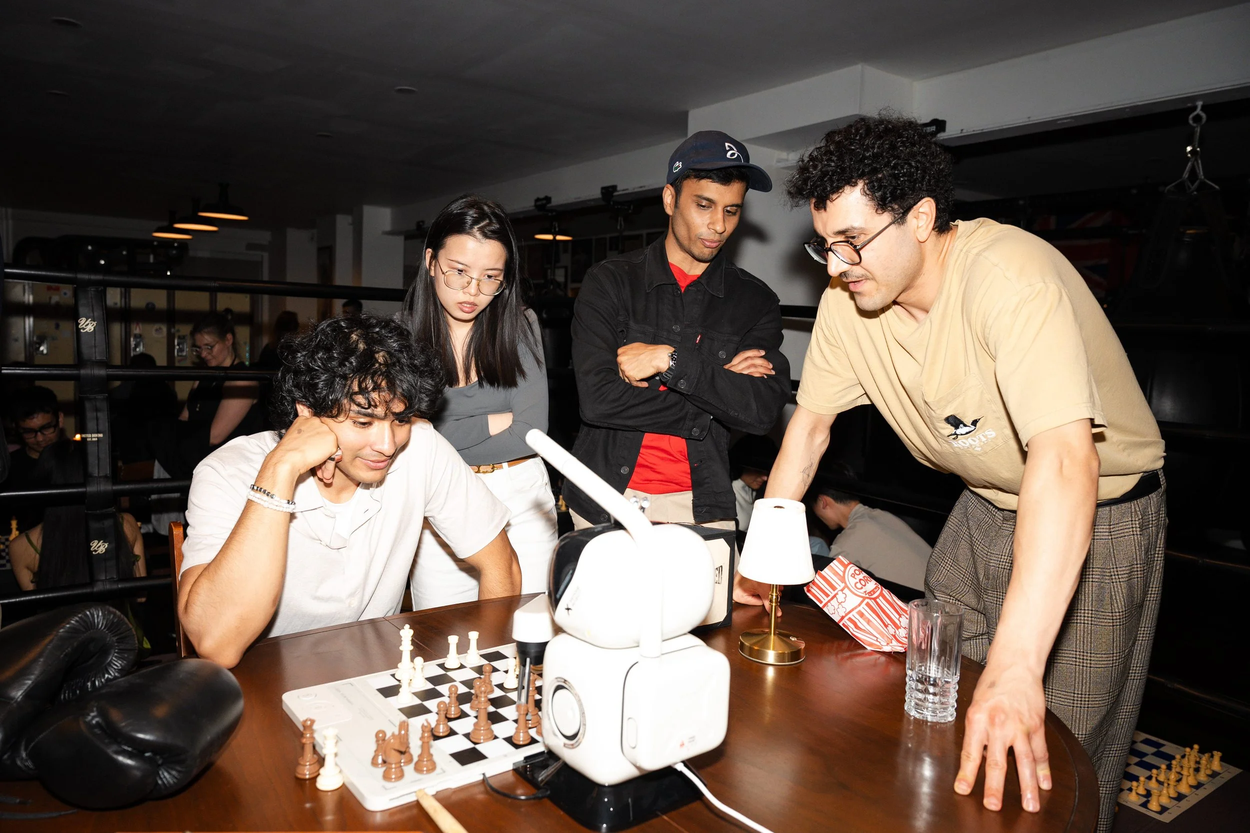A group of five people gathered around a table playing chess in a room with dim lighting. One person is seated with hand on head, another is standing pointing at the chessboard, the others are watching.