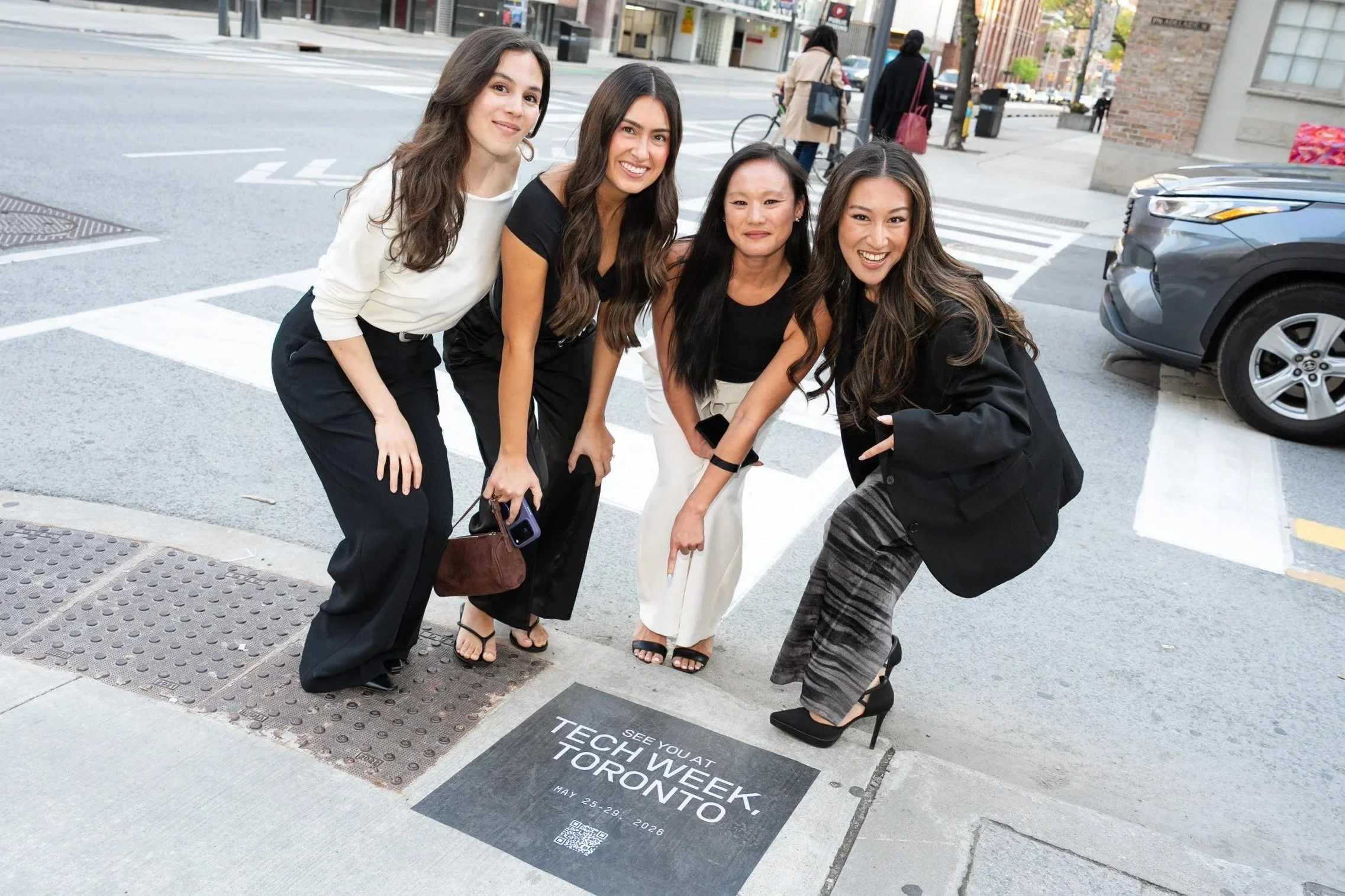 Four women smiling and posing on a city crosswalk, with a promotional ground sticker for the "Tech Week Toronto" event visible at their feet, in an urban street setting.