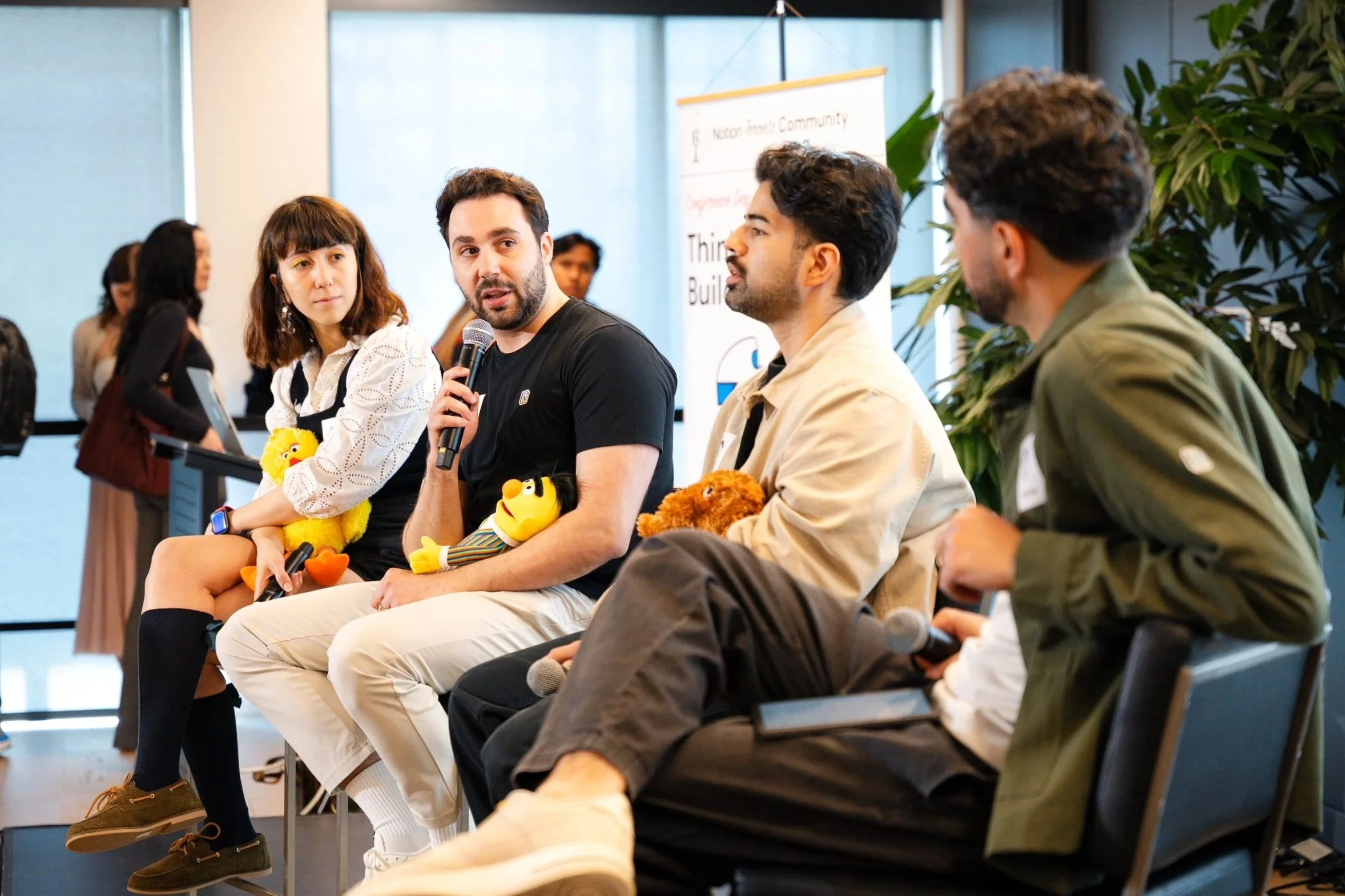 Group of four people sitting on chairs, participating in a panel discussion at an indoor event, with a woman standing and a banner in the background.