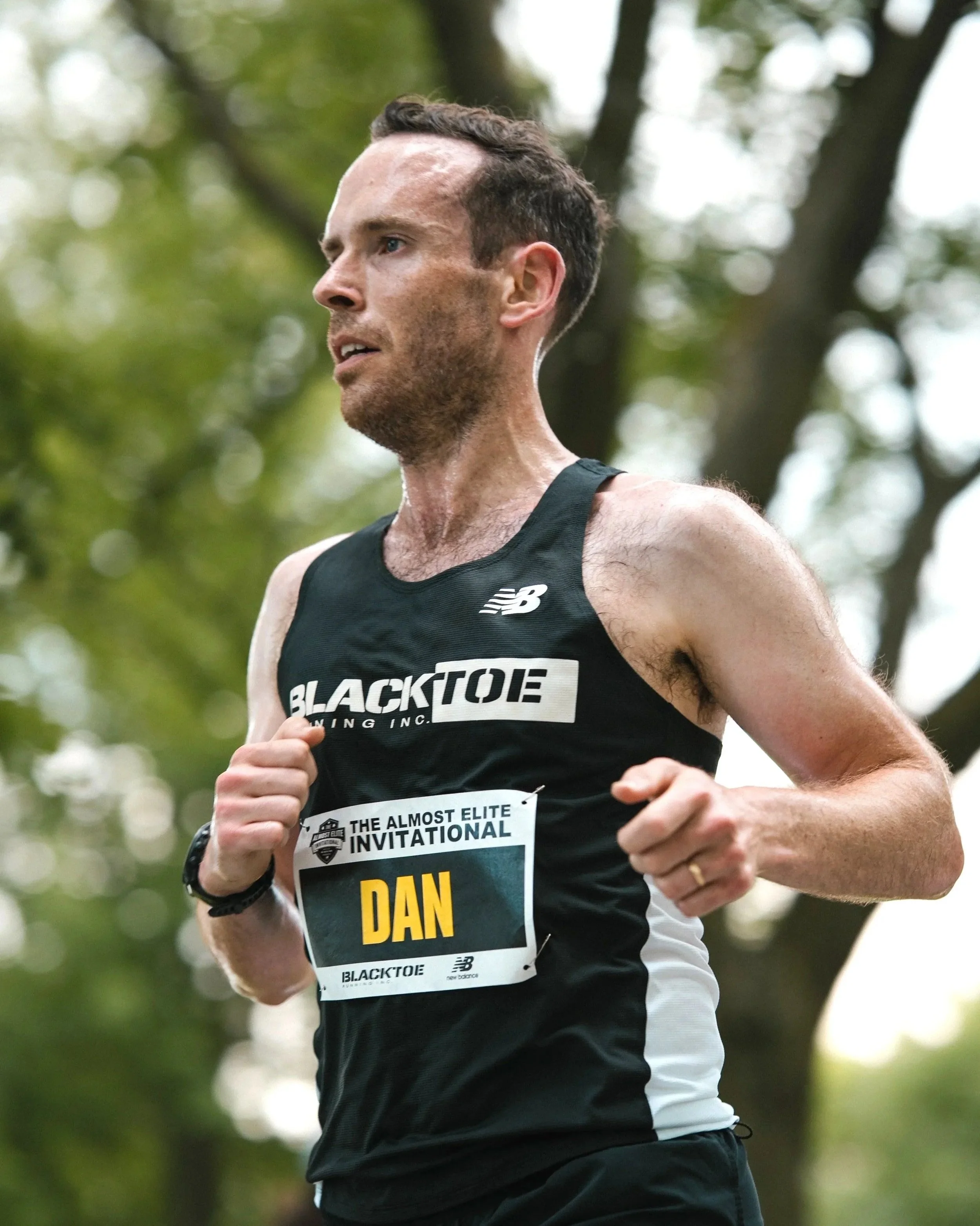 A male runner with short dark hair, wearing a black Running Inc. tank top, running through a park with trees and green foliage, and a race bib on his chest.