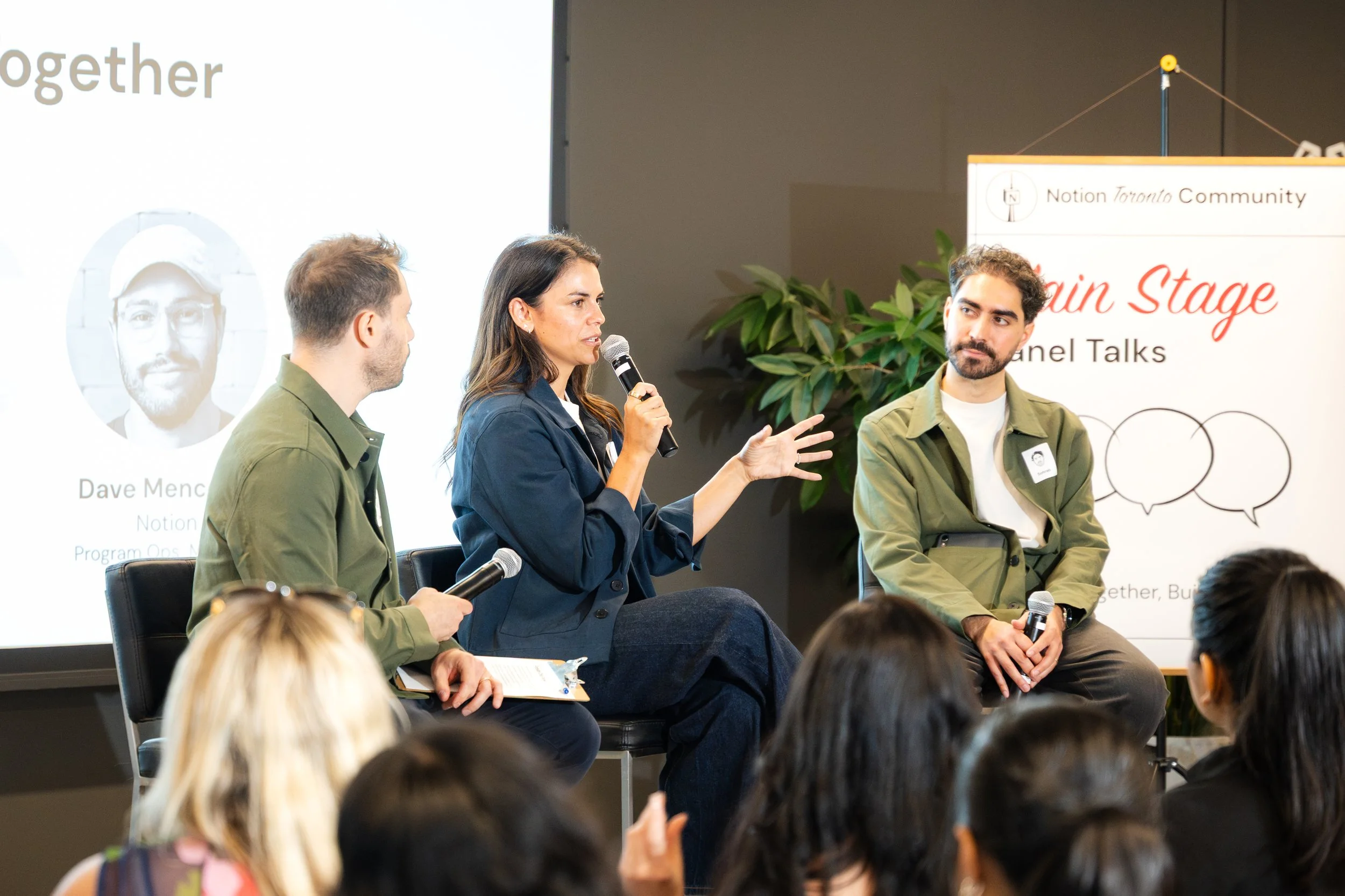 A woman speaking into a microphone during a panel discussion, flanked by two men holding microphones, with an audience in front. A screen behind them displays a partial presentation slide with an image of a man and some text.