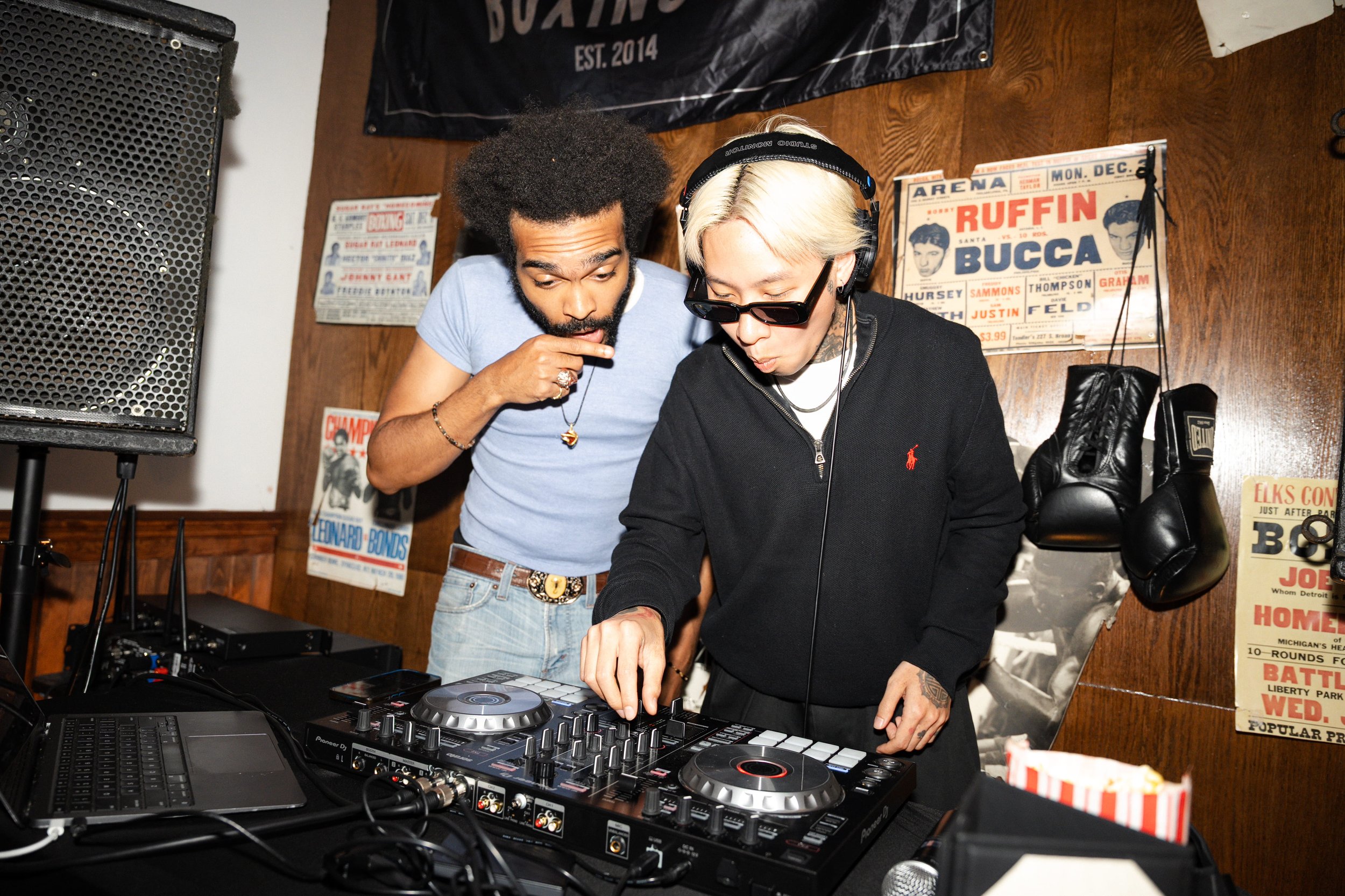 Two men are DJing and looking at a DJ controller. One is wearing sunglasses and headphones, and the other has an afro hairstyle and is wearing a light blue t-shirt.