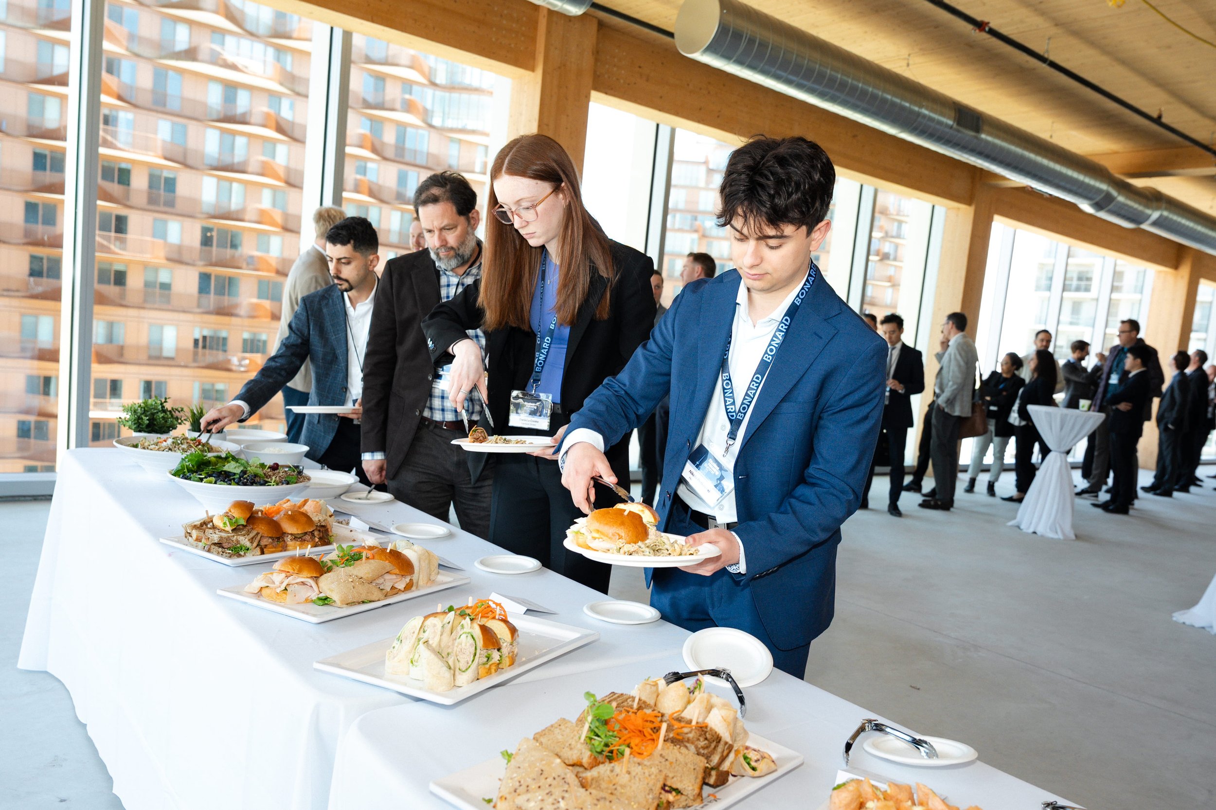 People at a catering buffet table with sandwiches and salads inside a modern building with large windows and a city view.