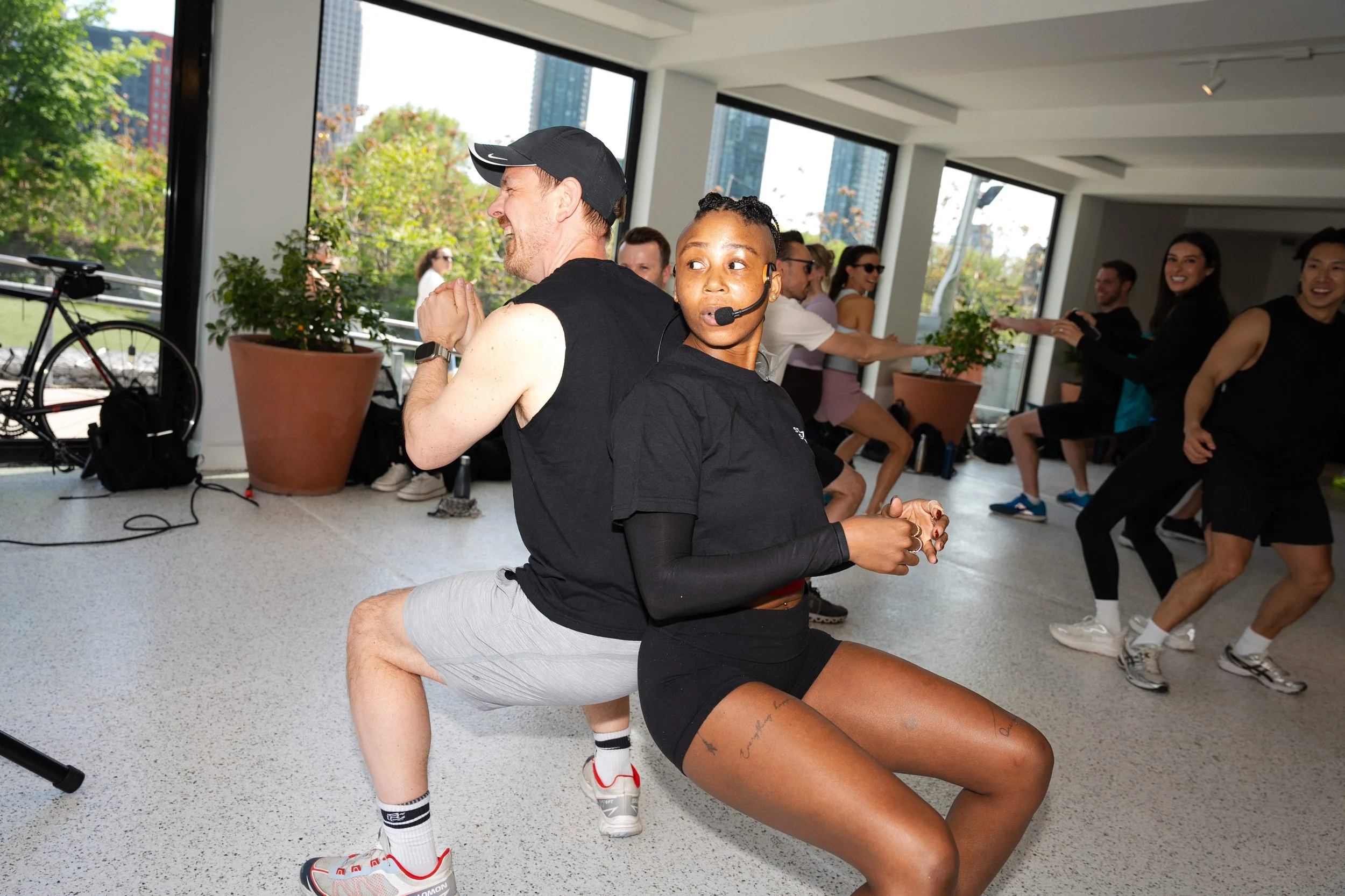 Two people exercising together in a fitness class, with a group smiling and stretching in the background, set inside a bright room with large windows and cityscape views.
