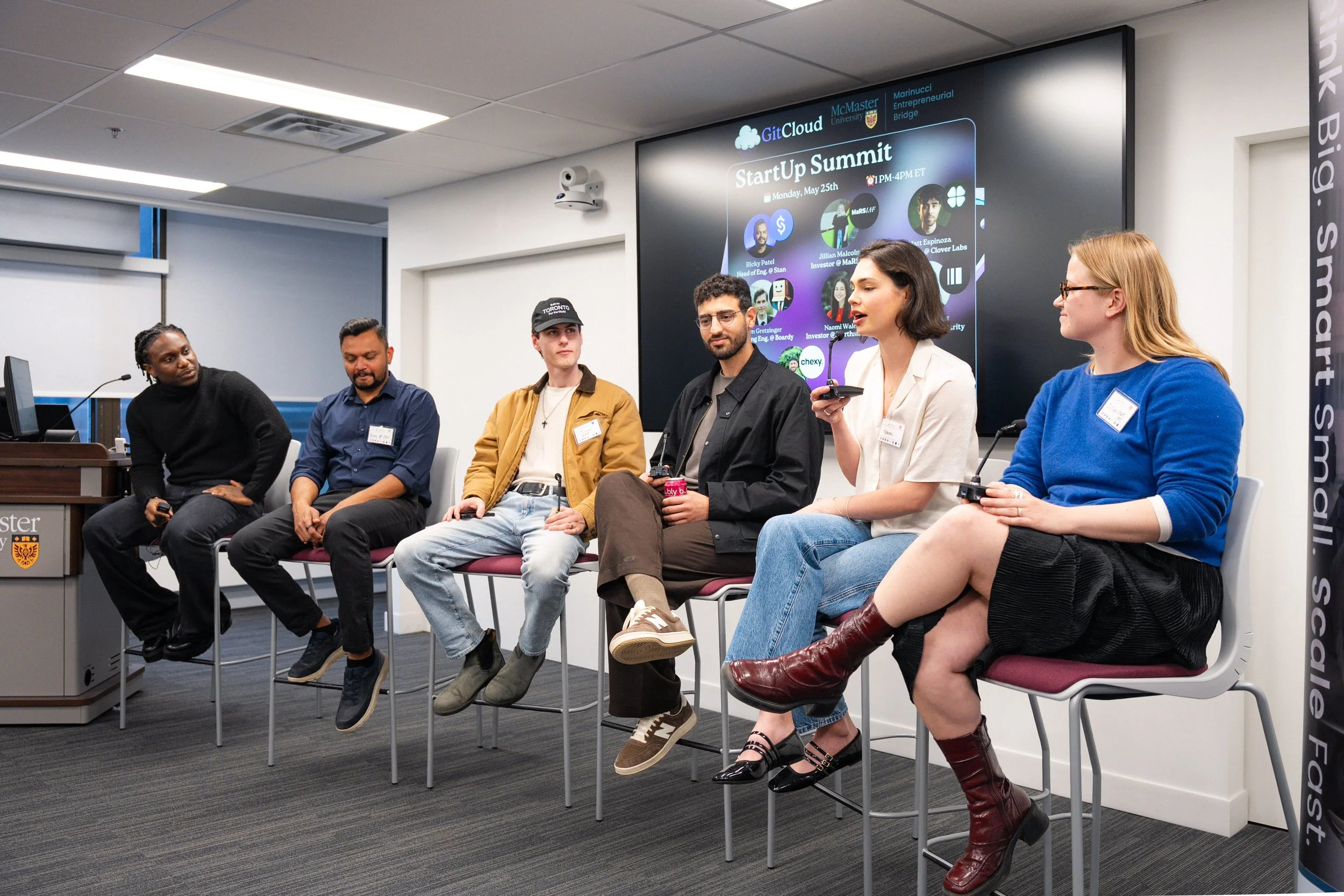 Six individuals seated on chairs at a panel discussion during a startup summit held at McMaster University, with a large screen displaying event details behind them.