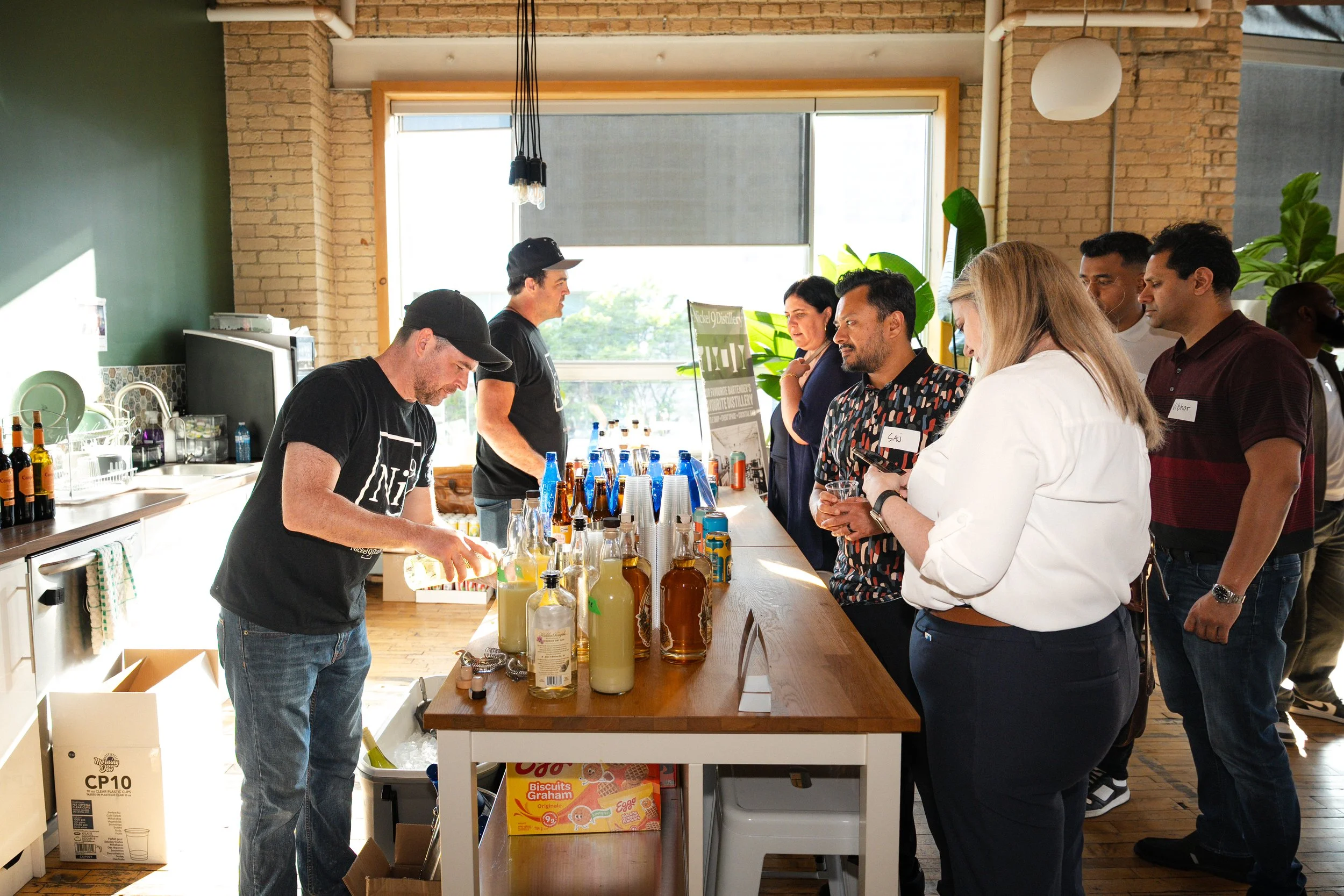 People attending a tasting event at a brewery or distillery, with bottles and tasting glasses on a wooden table, and staff serving drinks.