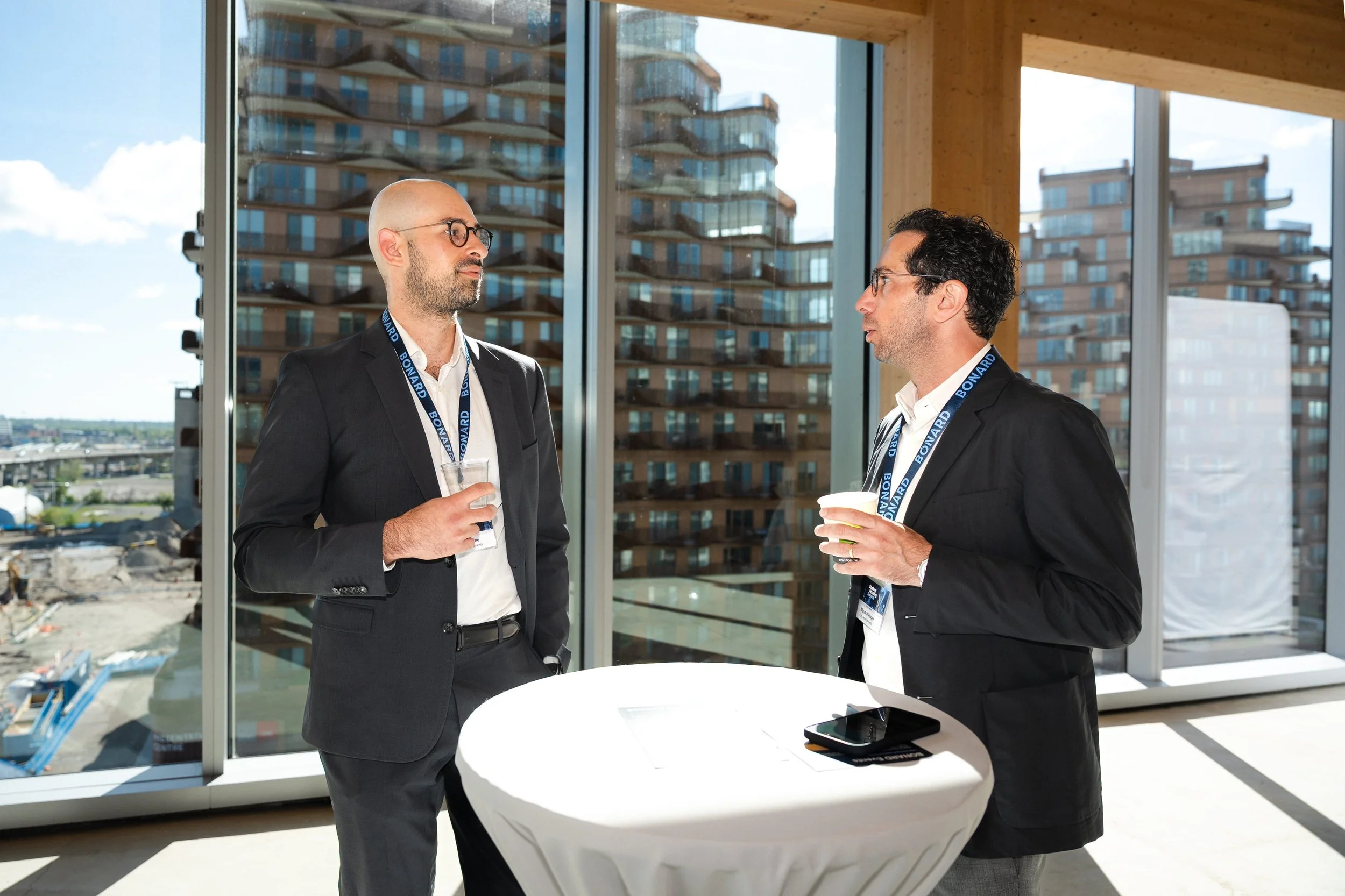 Two men in business suits and glasses having a conversation at a networking event inside a modern building with large windows and city views, each holding a drink, and a small round table with a smartphone and a flyer on it.