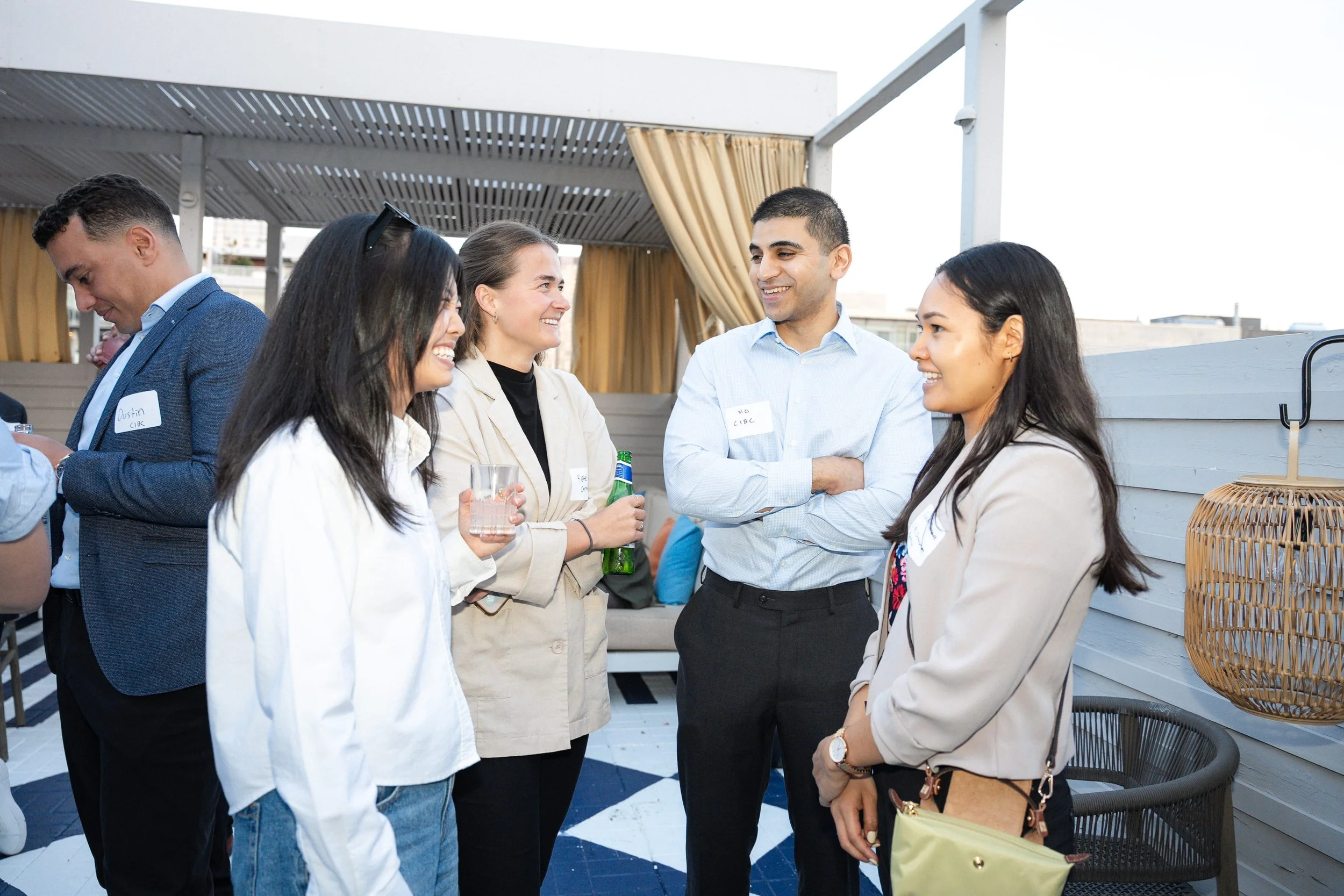 Group of five diverse professionals having a conversation at an outdoor rooftop event, some holding drinks, wearing business casual attire, with a modern setting and decorative elements in the background.