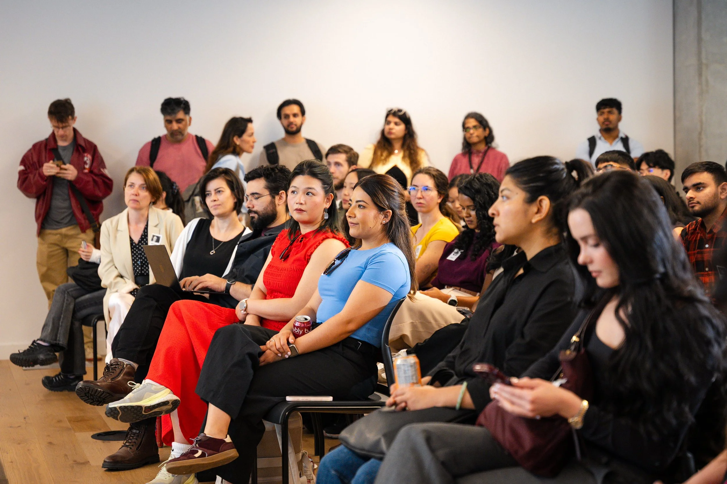 A diverse group of people sitting and standing in a room, attending an event or conference, with some using electronic devices and others listening or waiting.