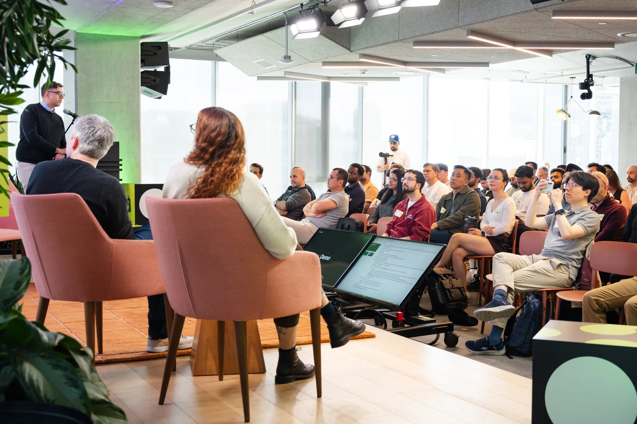 A speaker giving a presentation to an audience in a modern, well-lit conference room with floor-to-ceiling windows, while attendees listen and take notes.