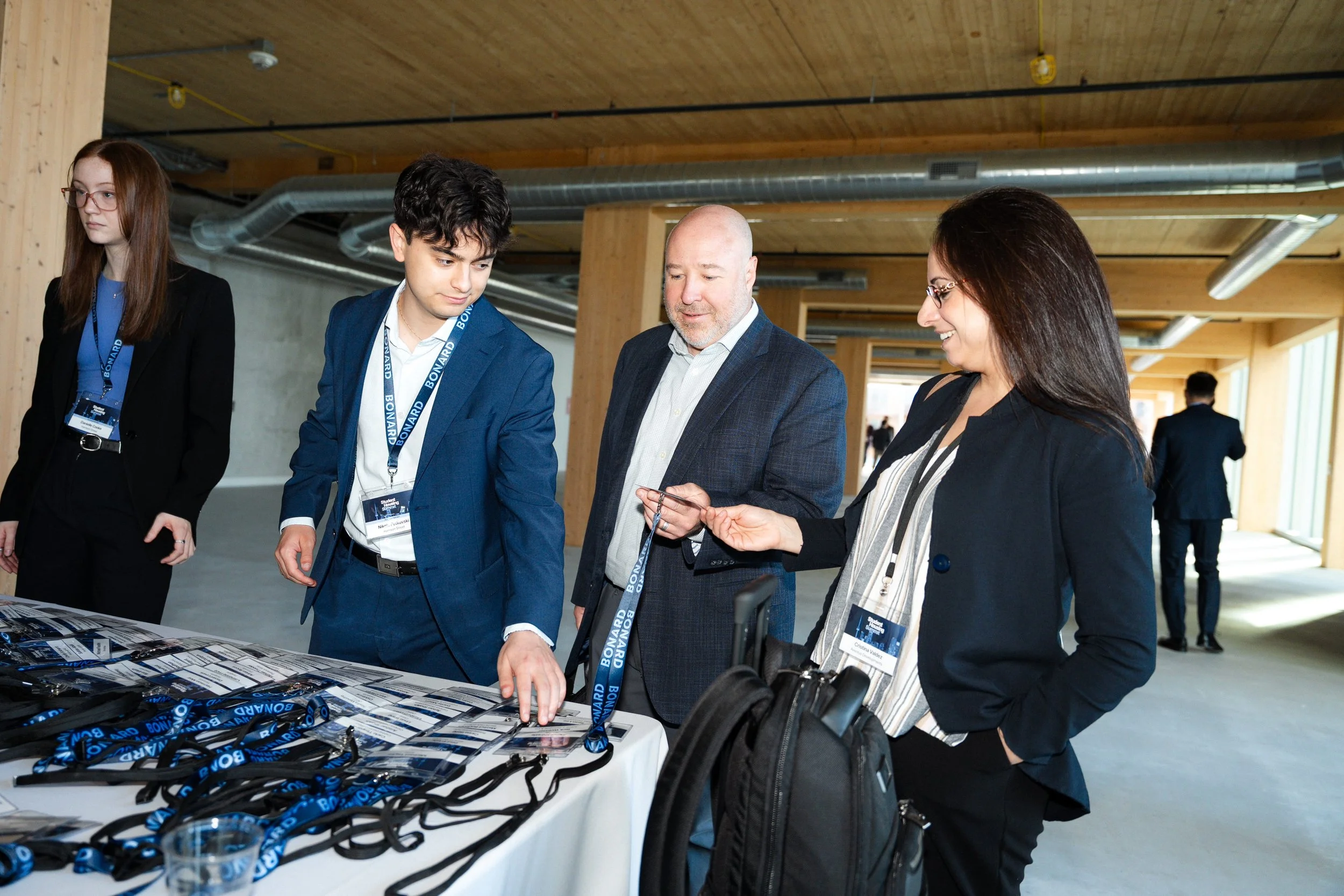 People at an event registration or check-in table, wearing name badges and lanyards, in a modern indoor venue with wooden ceiling and exposed ductwork.