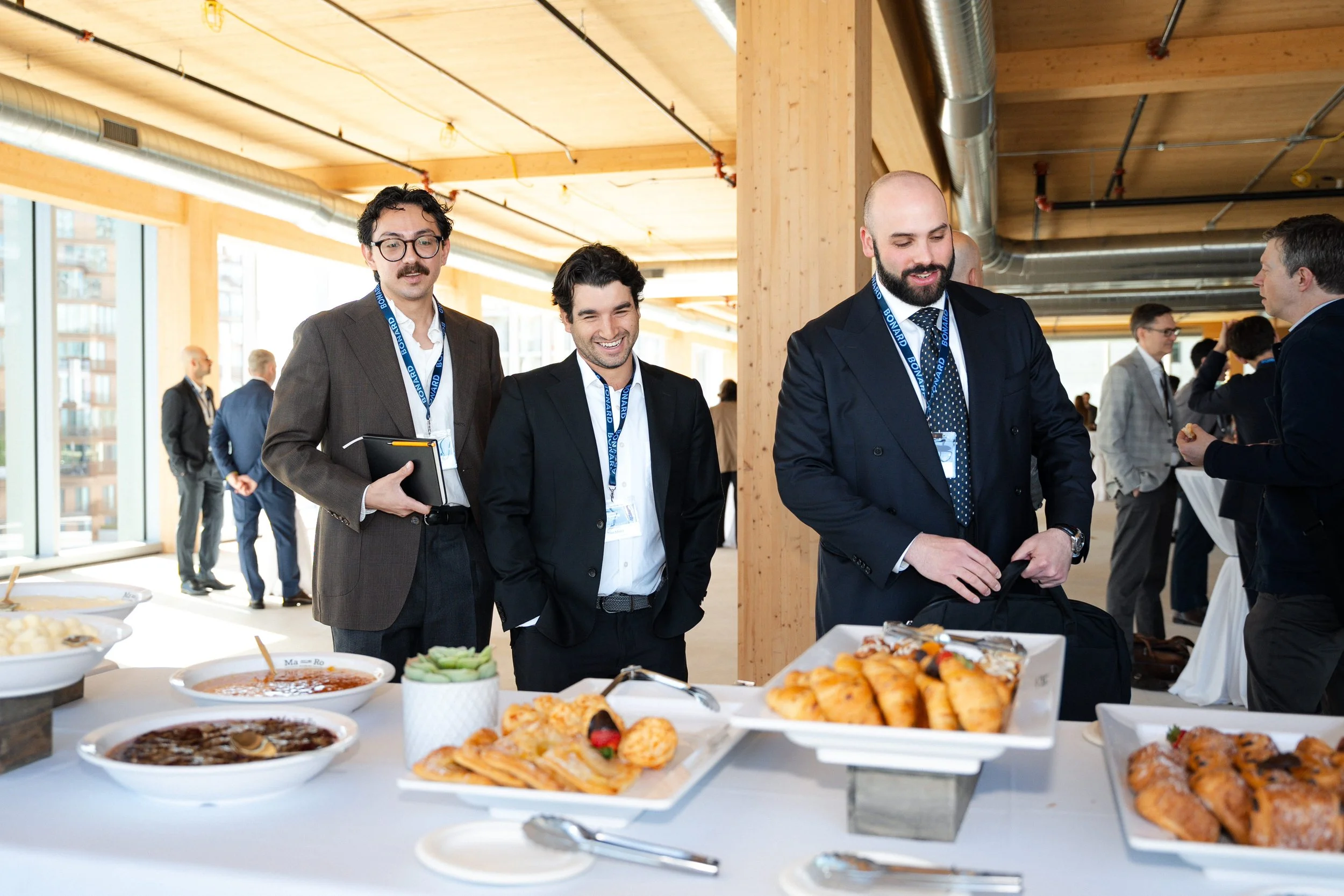 Business professionals at a buffet table with various dishes during a conference or event in a modern venue with wooden beams and large windows.