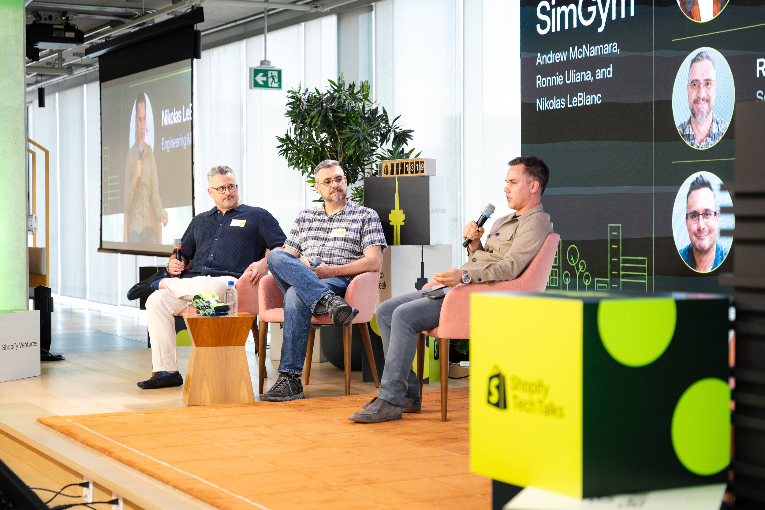 Three men sitting on stage during a panel discussion at Shopify Ventures event. One man is speaking into a microphone, while the others listen. Large screen behind them displays names and photos of panelists, including Andrew McNamara, Ronnie Uliana,