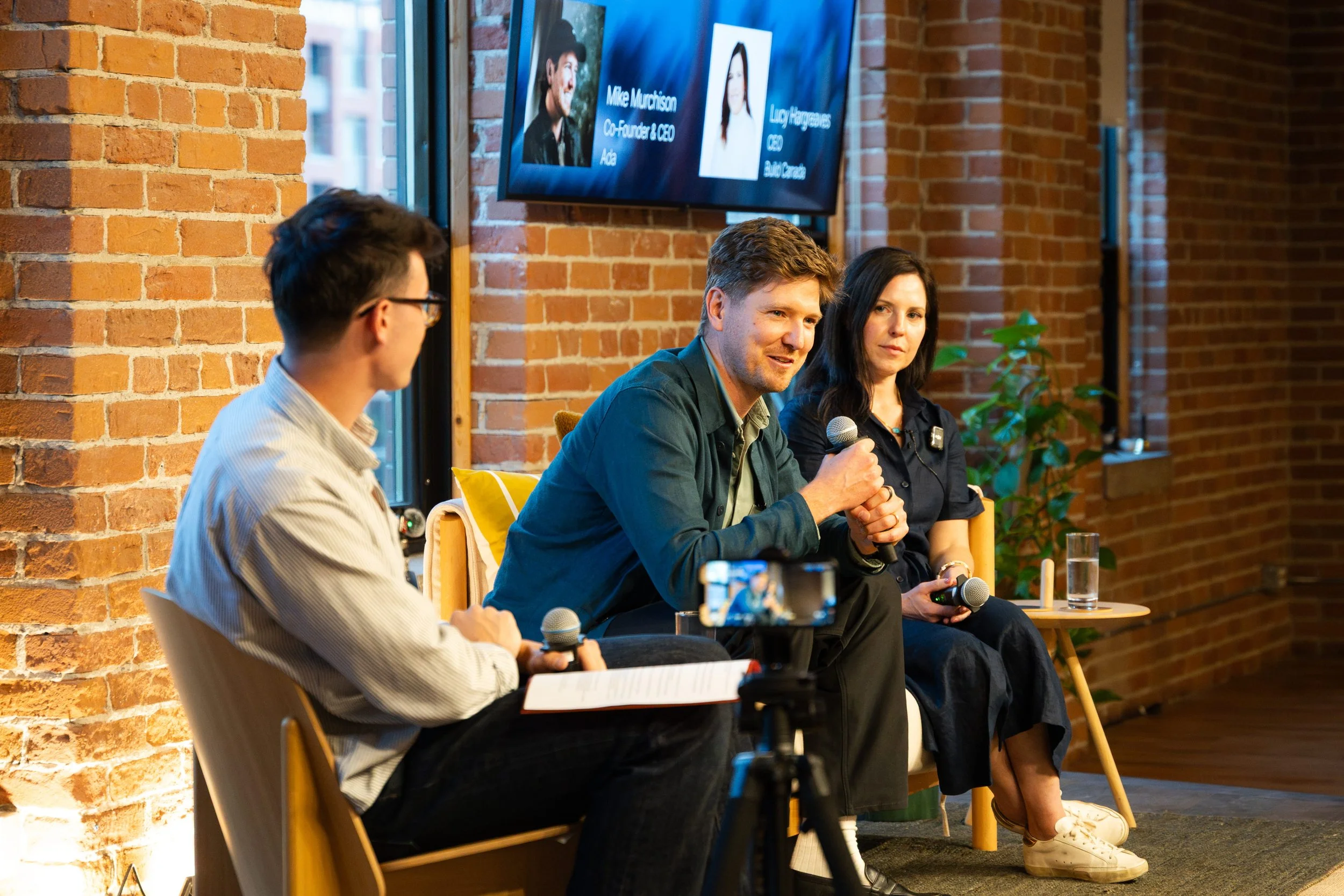 A panel of three people sitting in a row in front of a brick wall, participating in a discussion at an event. One person is speaking into a microphone, while the others listen. A screen behind them displays photos and names of the panelists.