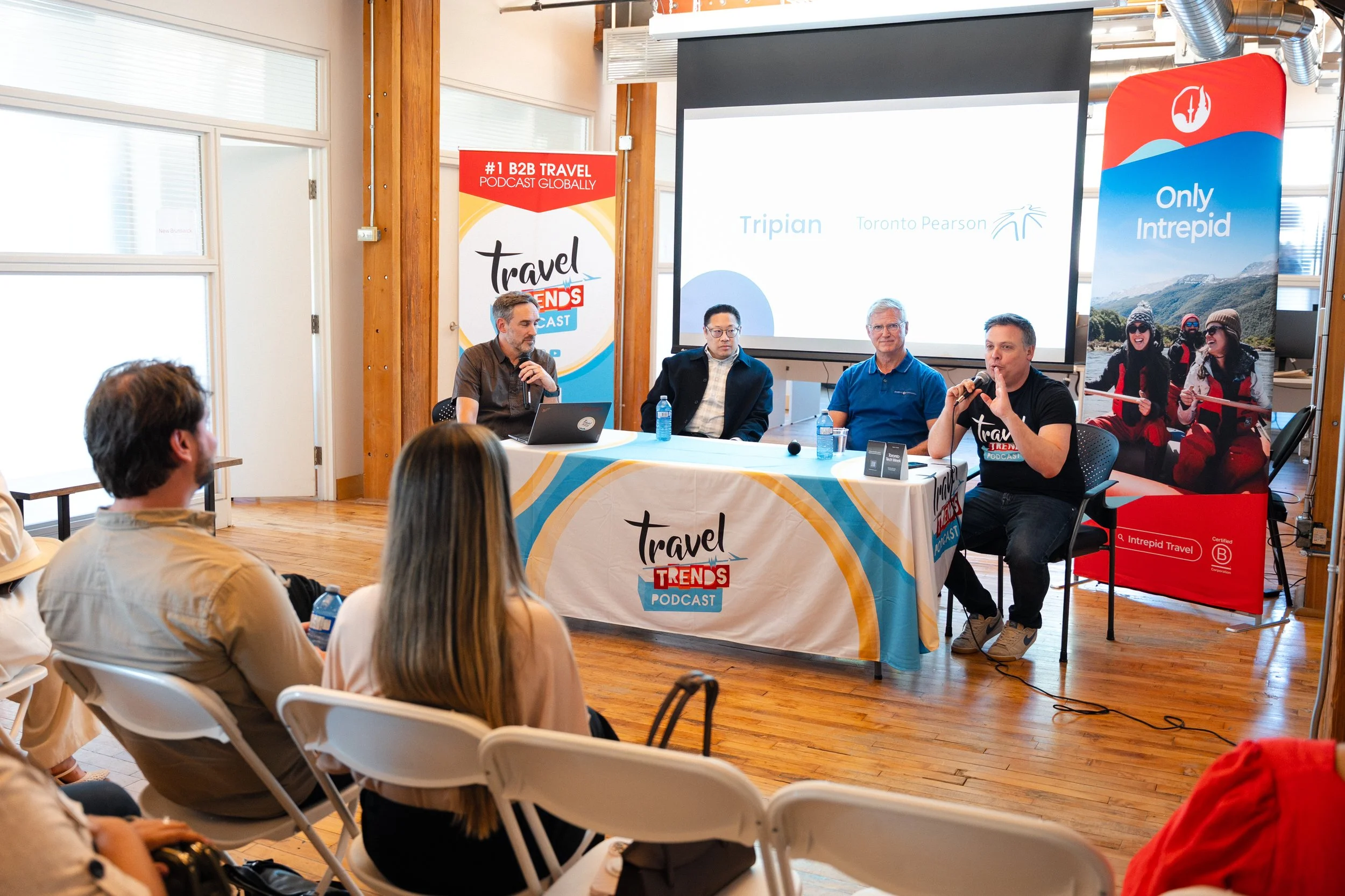Panel discussion at a travel podcast event with five men seated at a table, microphones, and a large screen displaying speaker names, with promotional banners for 'Travel Trends Podcast' and 'Only Intrepid' in the background.