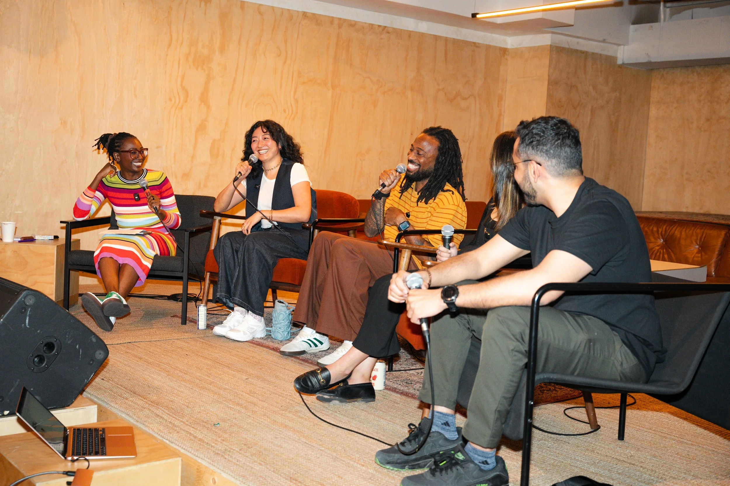 A diverse group of five people sitting on a stage, engaged in a panel discussion. They are holding microphones and smiling. There is a laptop and a speaker in front of them, and the background features plain, light-colored walls.