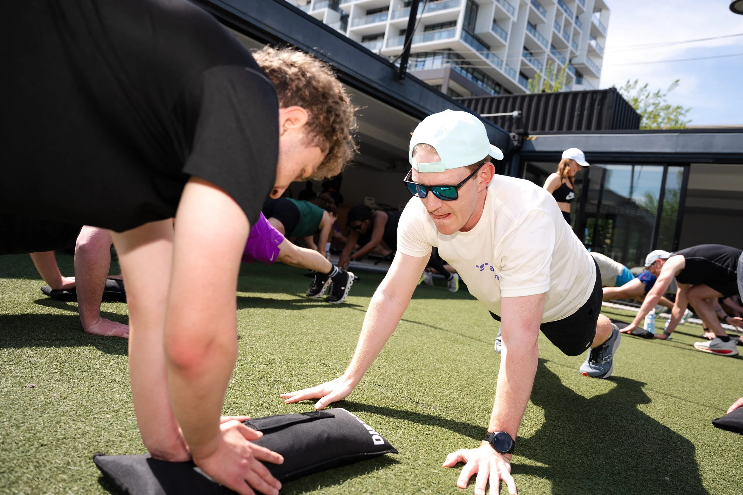 People participating in an outdoor yoga or fitness class on a green artificial turf, doing forearm and hand stretches, with a modern building and a partly cloudy sky in the background.