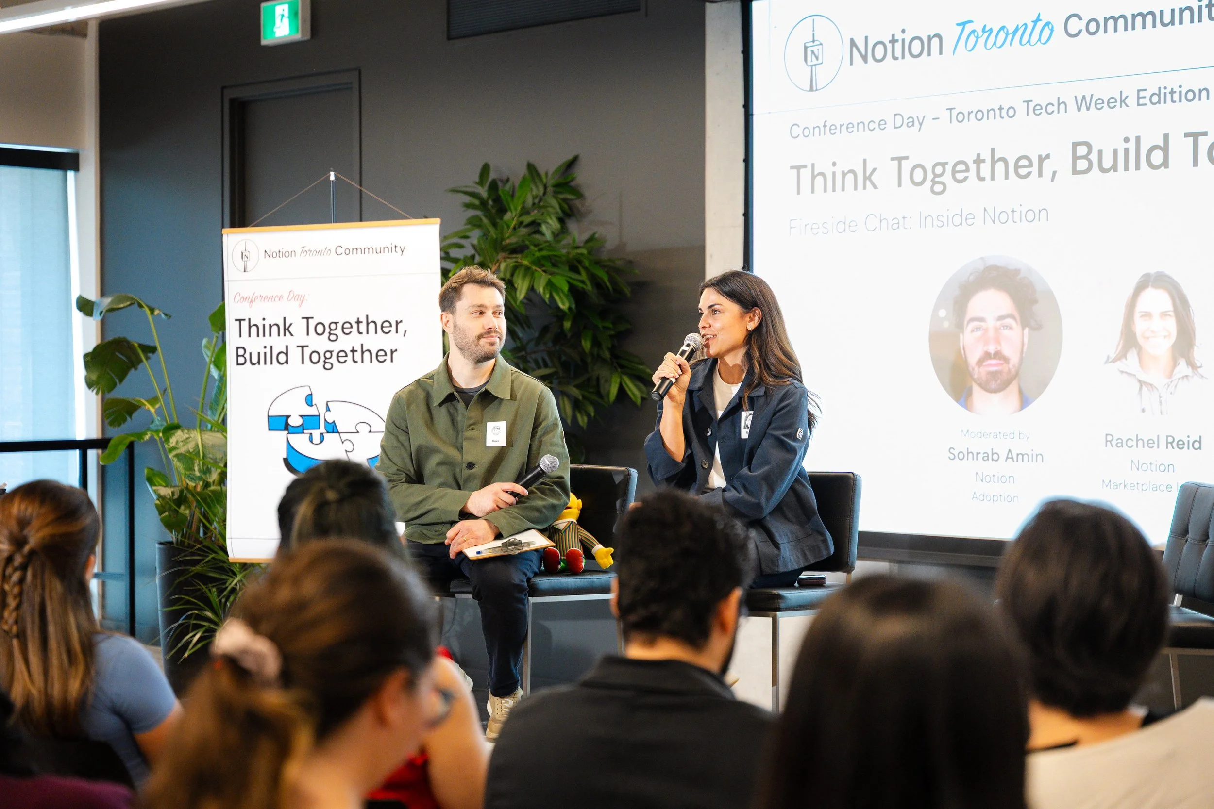 A woman speaks into a microphone while a man sits beside her, both on a stage at a conference with a large screen behind them displaying event information. The audience is visible in the foreground, and there are indoor plants and conference banners 