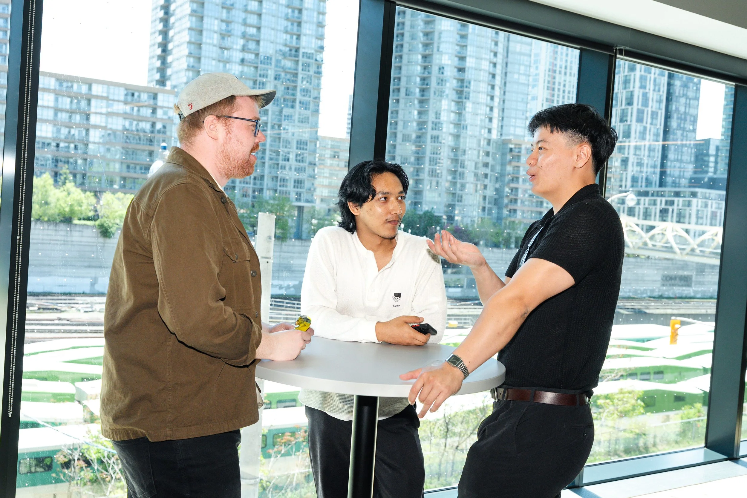 Three men engaged in conversation around a high table inside a modern building with large glass windows and city skyscrapers in the background.
