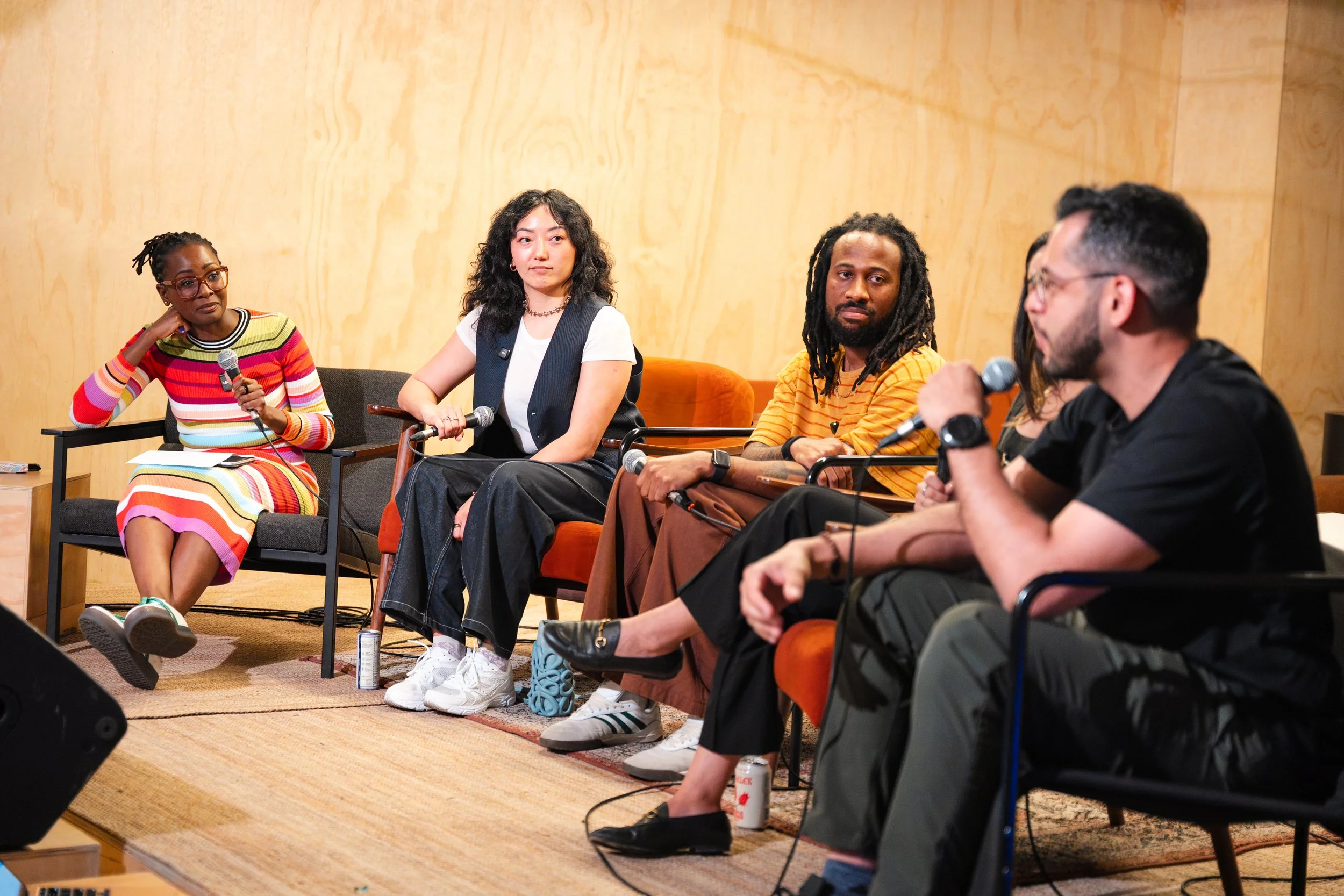 A diverse group of five people sitting in a panel discussion with microphones, a woman in a colorful striped dress holding a microphone, and others listening attentively against a plain wooden wall.