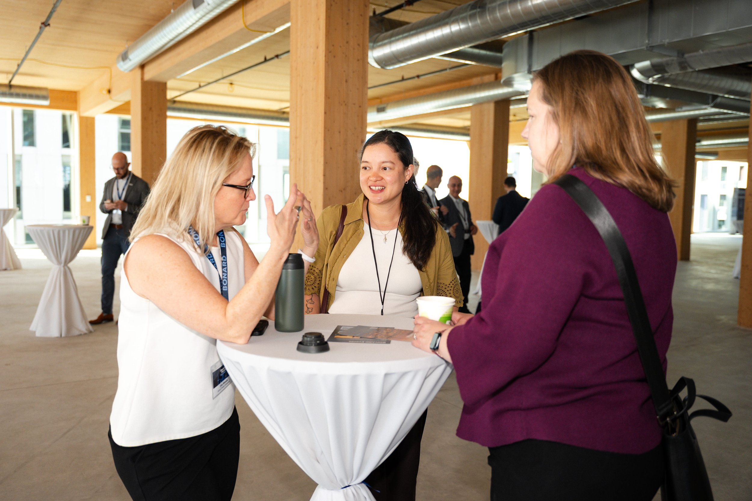 Three women standing around a tall, round table engaging in conversation at an indoor conference or networking event. One woman with blonde hair and glasses is speaking and gesturing with her hands, while another woman with dark hair, dressed in a ye