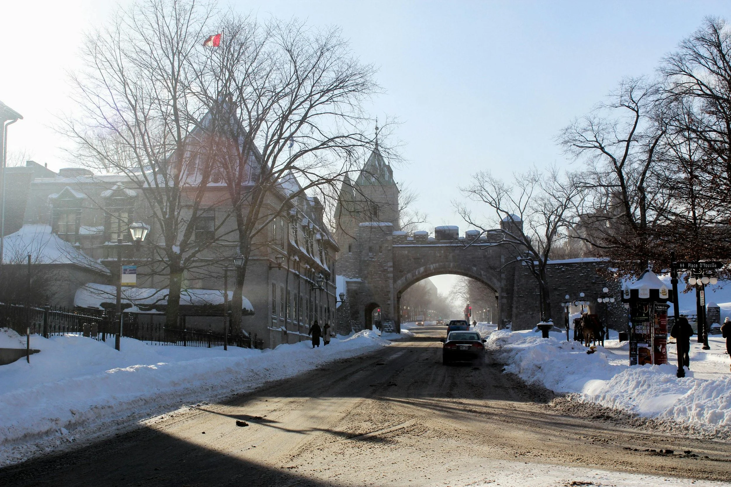 Rue enneigée avec un pont en pierre et une tour au fond, arbres sans feuilles, bâtiments anciens, quelques passants et voitures sous un ciel clair hivernal.