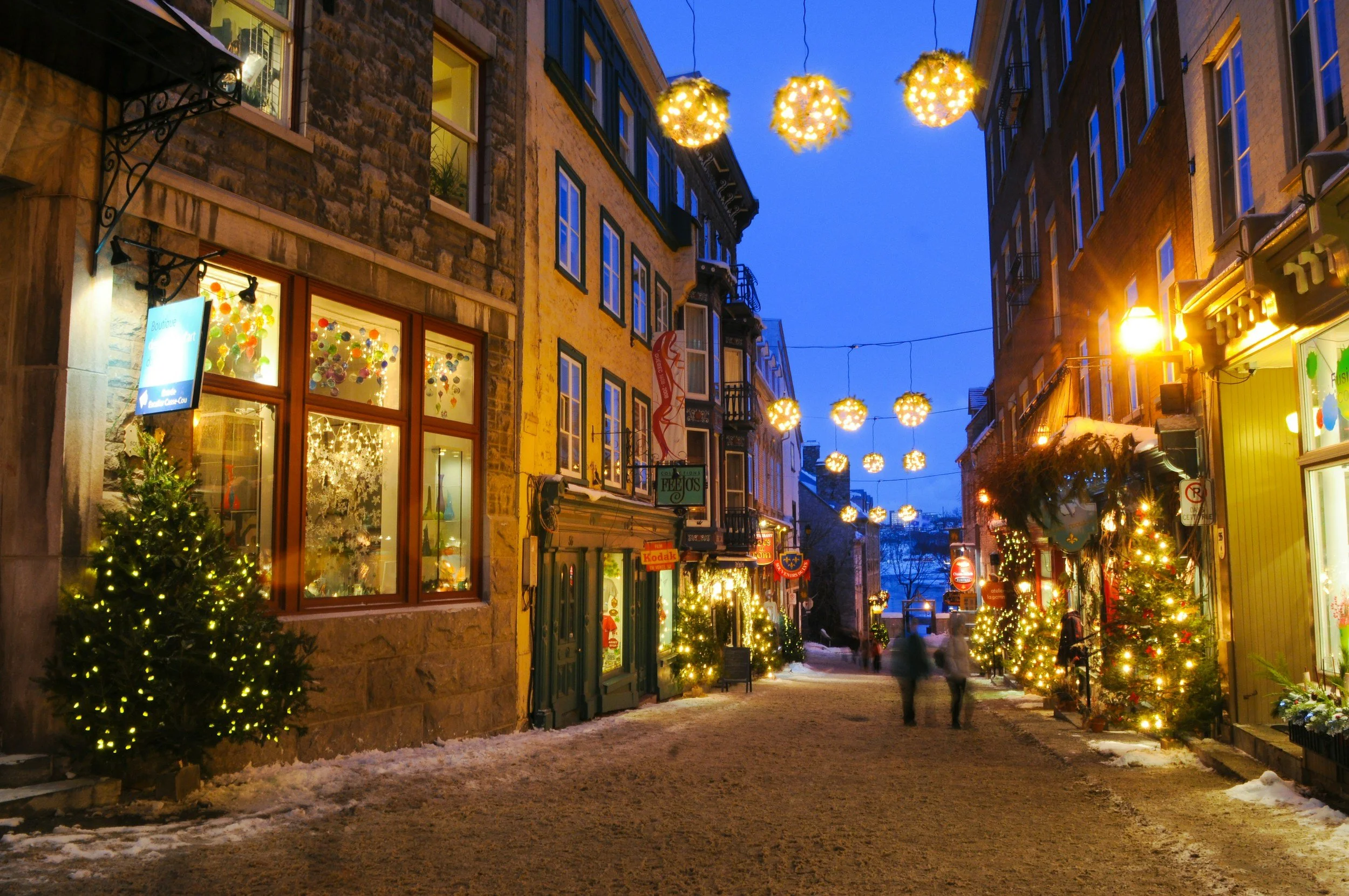 Une rue piétonne décorée pour Noël avec des sapins éclairés, des guirlandes lumineuses suspendues, et des boutiques illuminées, en soirée d'hiver avec la neige au sol.