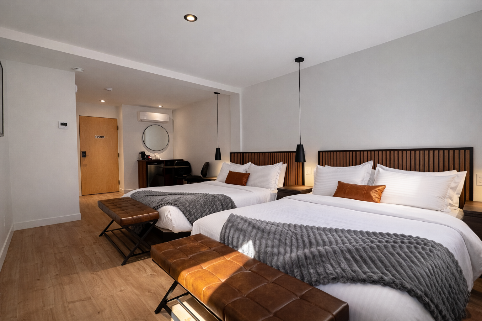 Deux grands lits dans une chambre d'hôtel moderne avec tête de lit en bois, couvertures gris foncé, oreillers blancs, lampes suspendues noires, bancs en cuir au pied des lits, et un coin bureau avec miroir rond en arrière-plan.